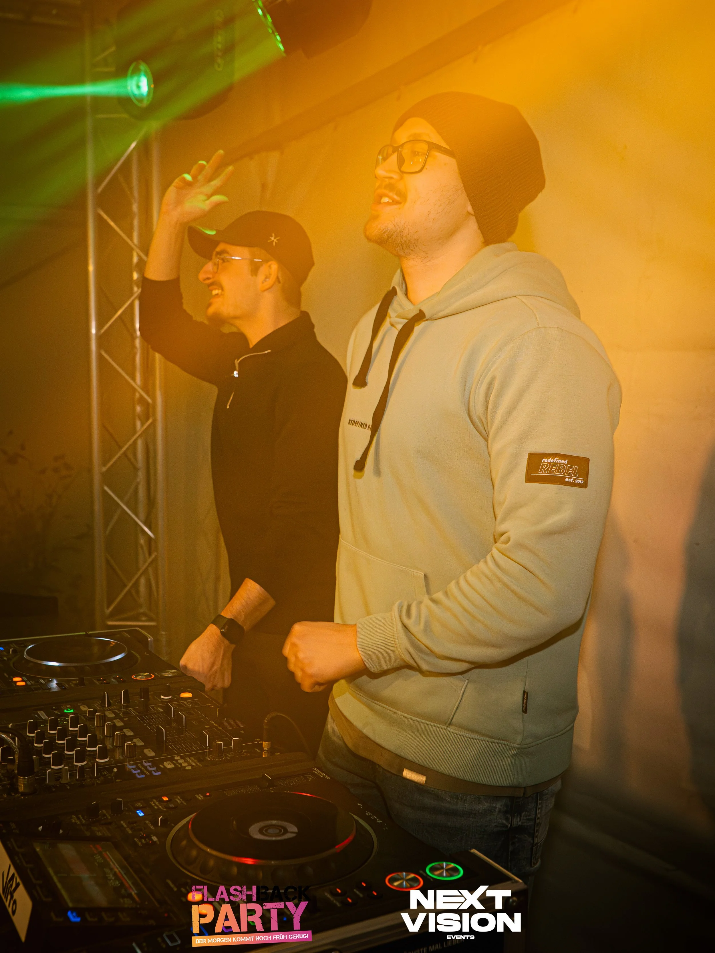 Zwei junge Männer am DJ-Deck bei einer Party, lachen und feiern, im Hintergrund bunte Lichtstrahlen.