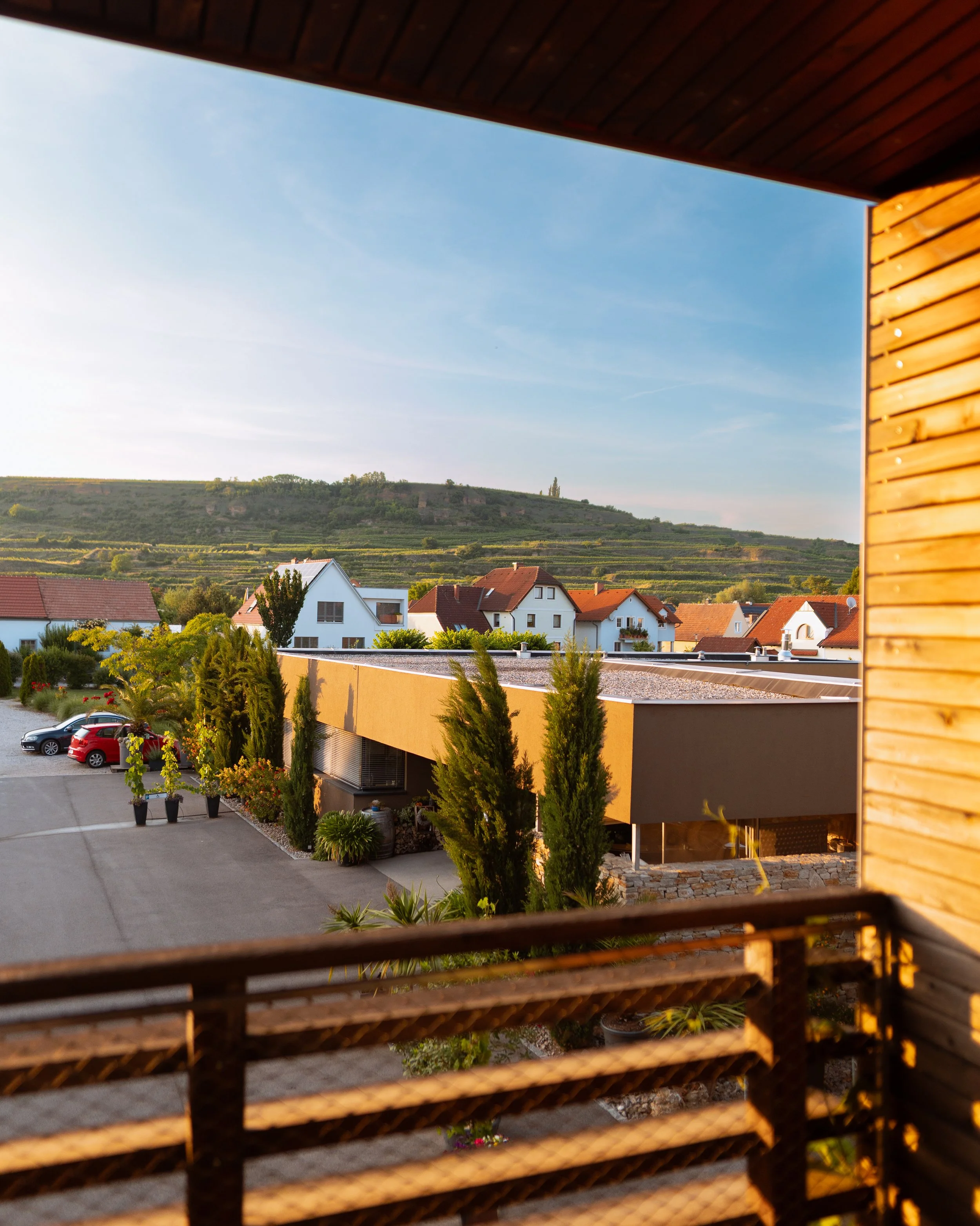 Blick von einem Balkon auf eine Wohnsiedlung mit Häusern, Bäumen und einem Hügel im Hintergrund bei Sonnenuntergang.