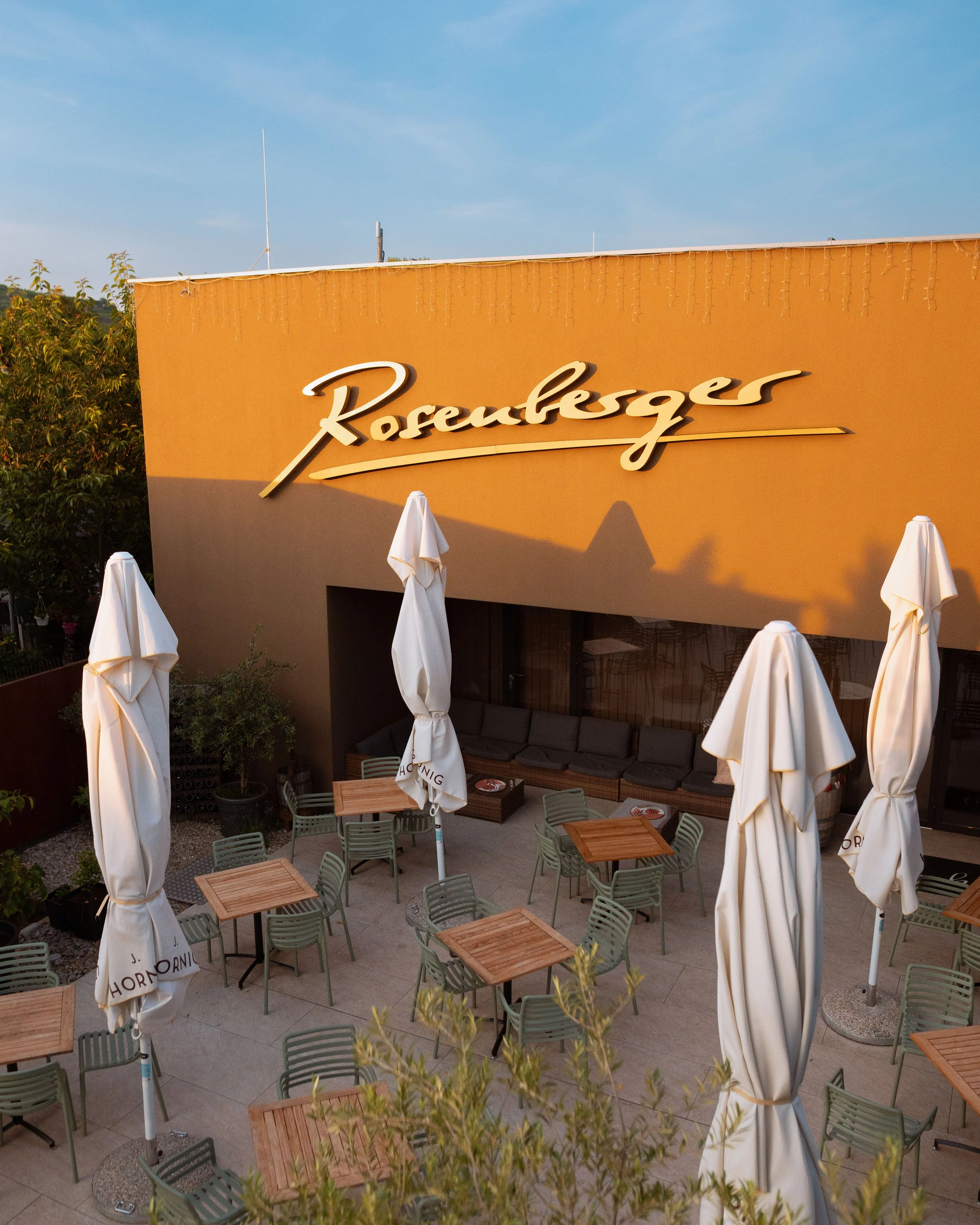 Außenansicht eines Restaurants mit dem Schriftzug 'Rosenberger' an der Wand, Tische mit Stühlen und geschlossenen Sonnenschirmen auf Terrasse bei Sonnenuntergang.
