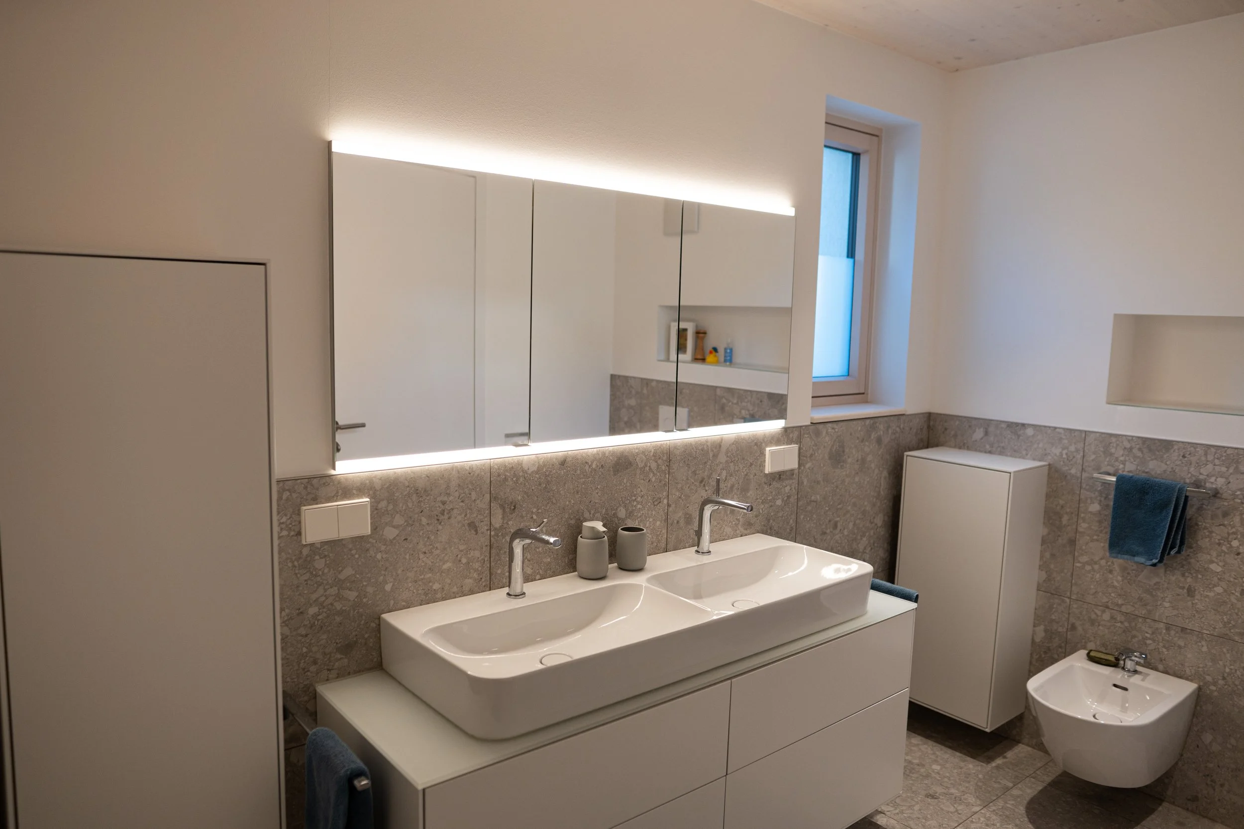 Modernes Badezimmer mit doppeltem Waschbecken, Spiegel mit integrierter Beleuchtung, Bidet, graue Fliesen, kleiner Schrank, blauer Handtuch, Fenster