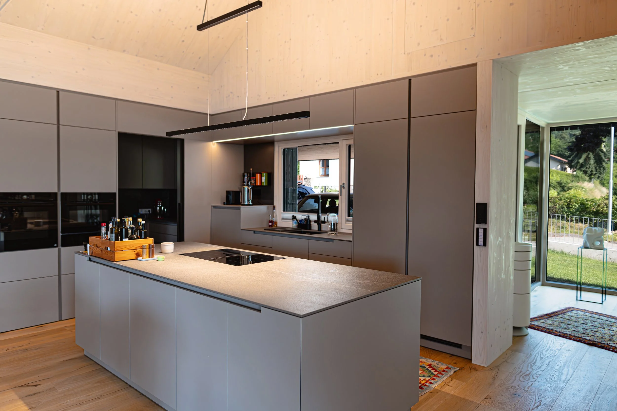 Moderne Küche mit grauen Schränken, einem Kücheninsel mit grauer Oberfläche, Herd, Wasserkocher, Geschirr und Dekorationen, große Fenster mit Blick auf den Garten, Holzdecke und Holzboden.