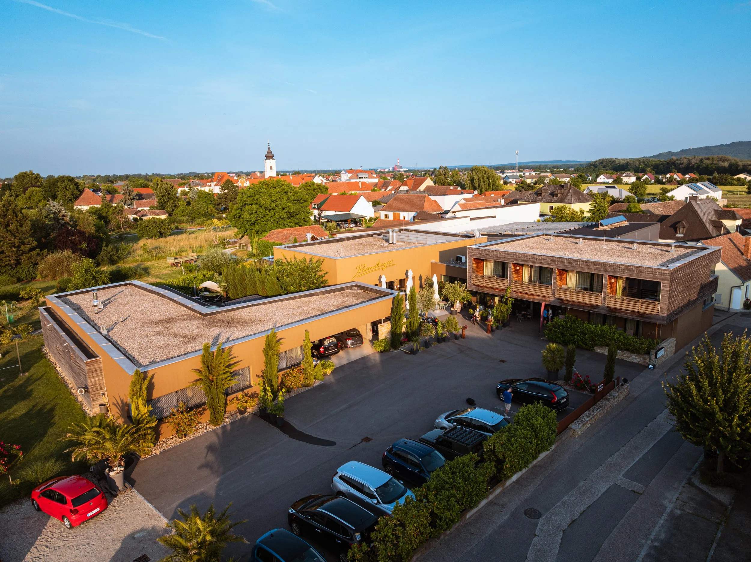 Vogelperspektive auf ein Hotelgebäude in einer ruhigen Stadt, umgeben von Bäumen und anderen Häusern, mit einem Parkplatz und einem klaren blauen Himmel.