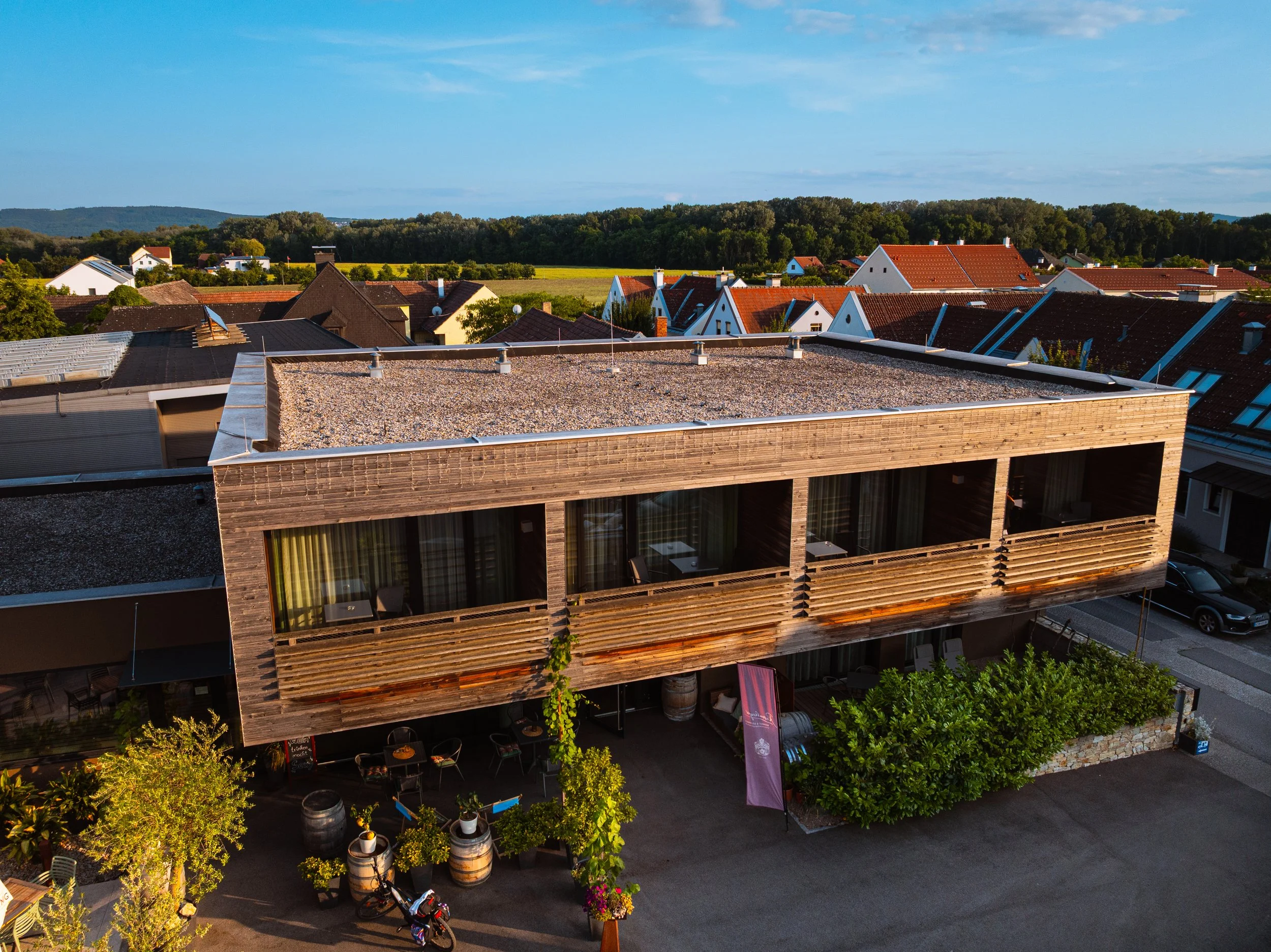 Ein modernes zweistöckiges Gebäude mit Holzfassade und großen Fenstern. Es hat ein Flachdach und befindet sich in einer Wohngegend mit anderen Häusern. Im Vordergrund sind einige Bäume, Pflanzen und eine Sitzmöglichkeit. Fahrräder sind zu sehen, und 