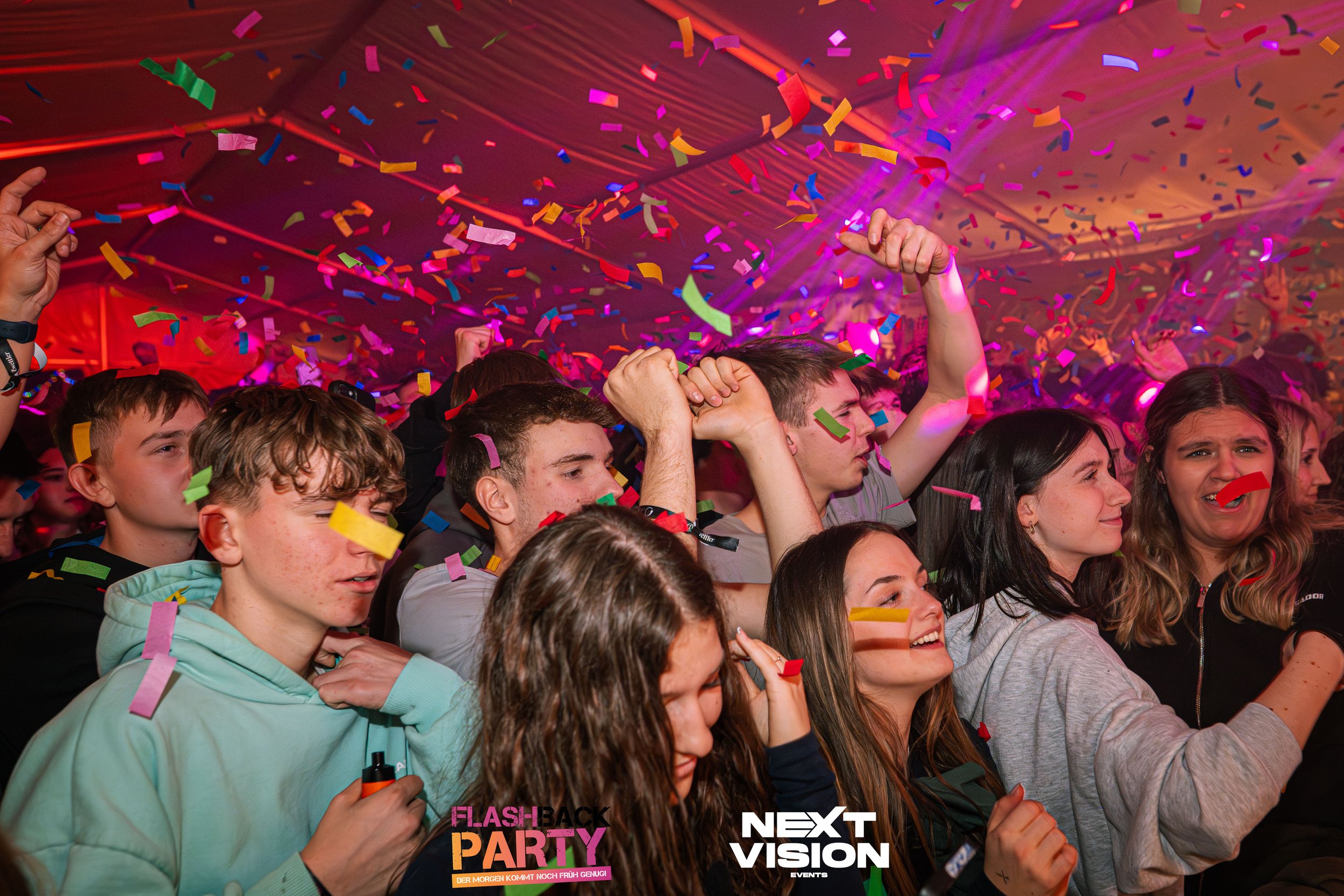 Junge Menschen feiern auf einer Party mit bunten Confettiregen, einige sind am Tanzen und einige scheinen begeistert, in einer festlichen Atmosphäre mit bunten Lichtern.