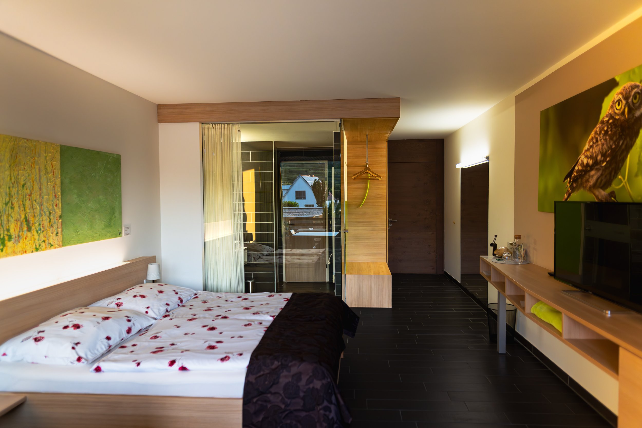 Ein modernes Hotelzimmer mit Doppelbett, Wandkunst, Flachbildfernseher, Holzmöbeln, offener Badezimmerbereich mit Dusche und Blick nach draußen auf einen Garten.