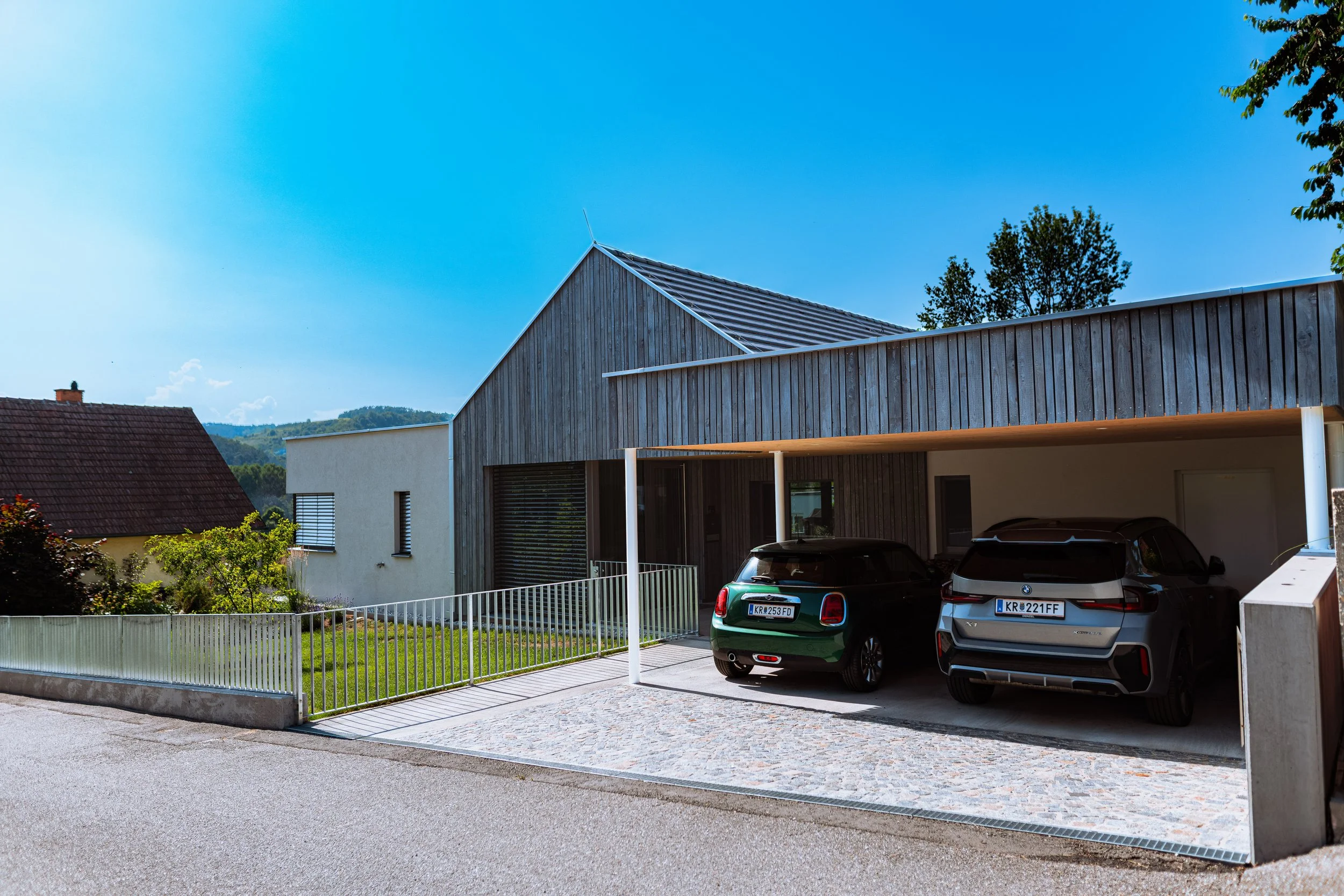 Moderne Einfahrt mit Carport, zwei Autos und einem Haus im Hintergrund auf einer sonnigen Straße in einer ländlichen Gegend.