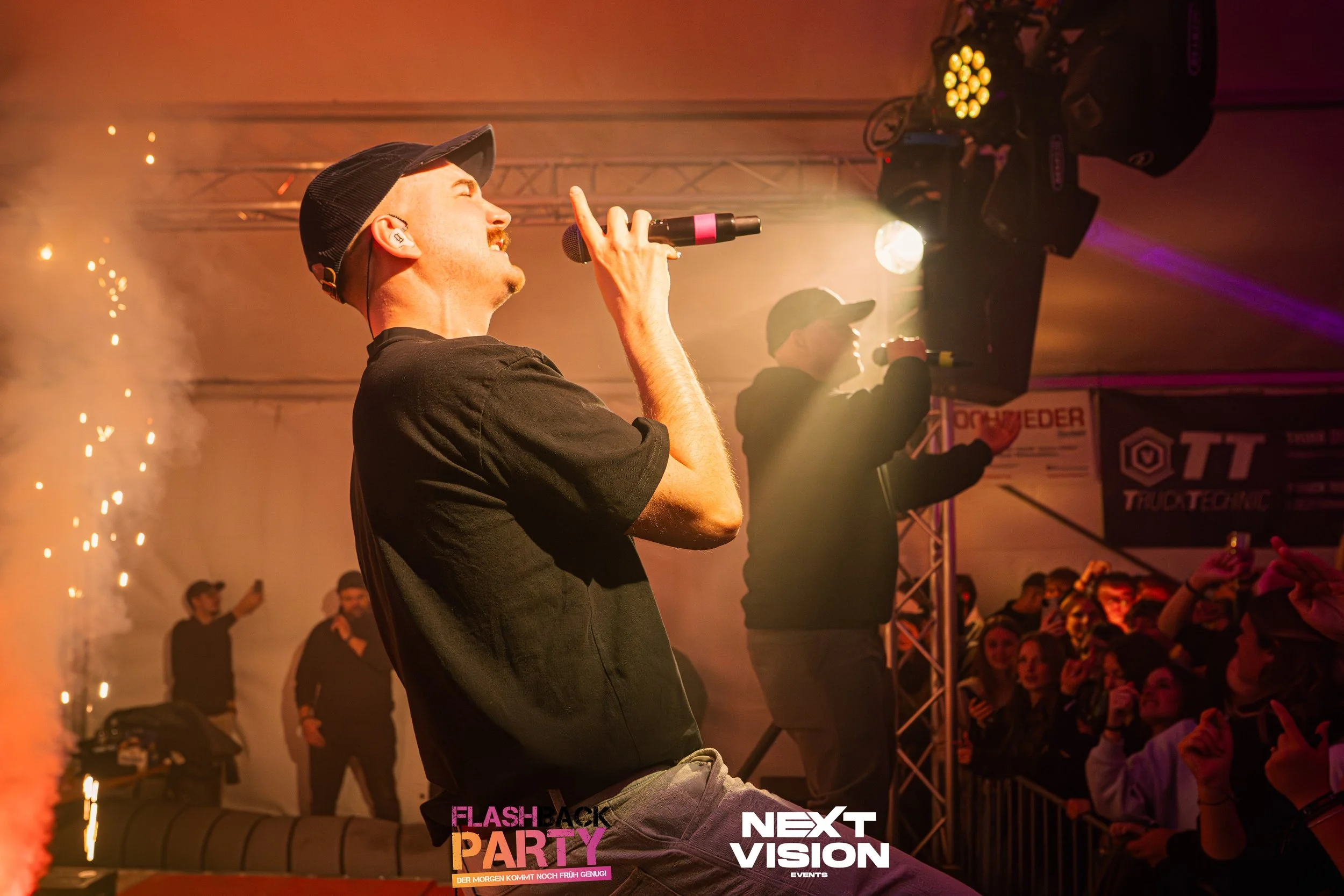 Ein Sänger bei einer Party im Inneren eines Zeltes, trägt eine schwarze Kappe und ein schwarzes T-Shirt, hält ein Mikrofon in der Hand und singt. Im Hintergrund sind weitere Personen und Publikum zu sehen, mit Bühnenlichtern und Effekten.