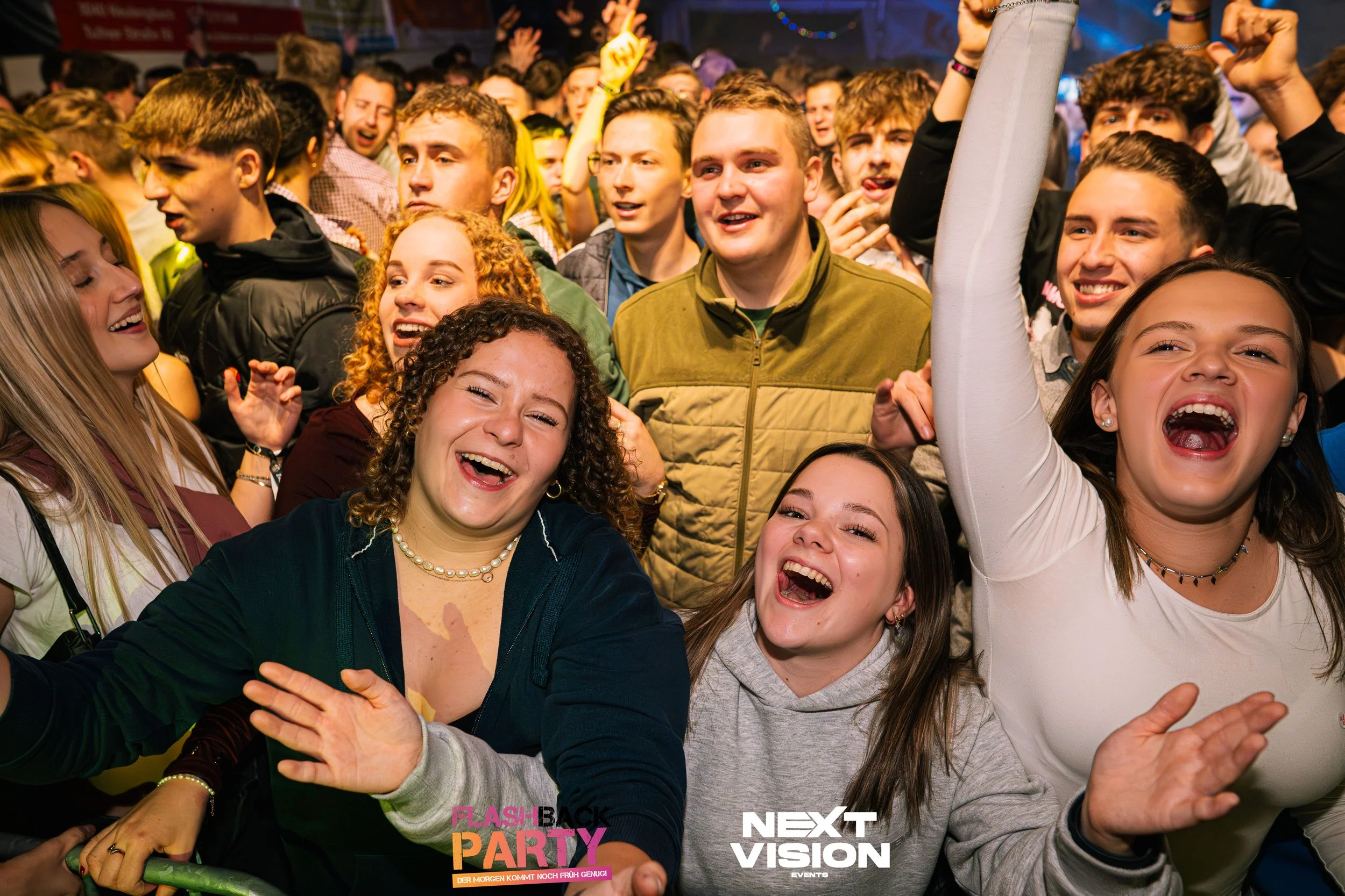 Menschen beim feierlichen Event, lachen und feiern auf der Bühne oder in der Menge, bunte Lichter im Hintergrund.