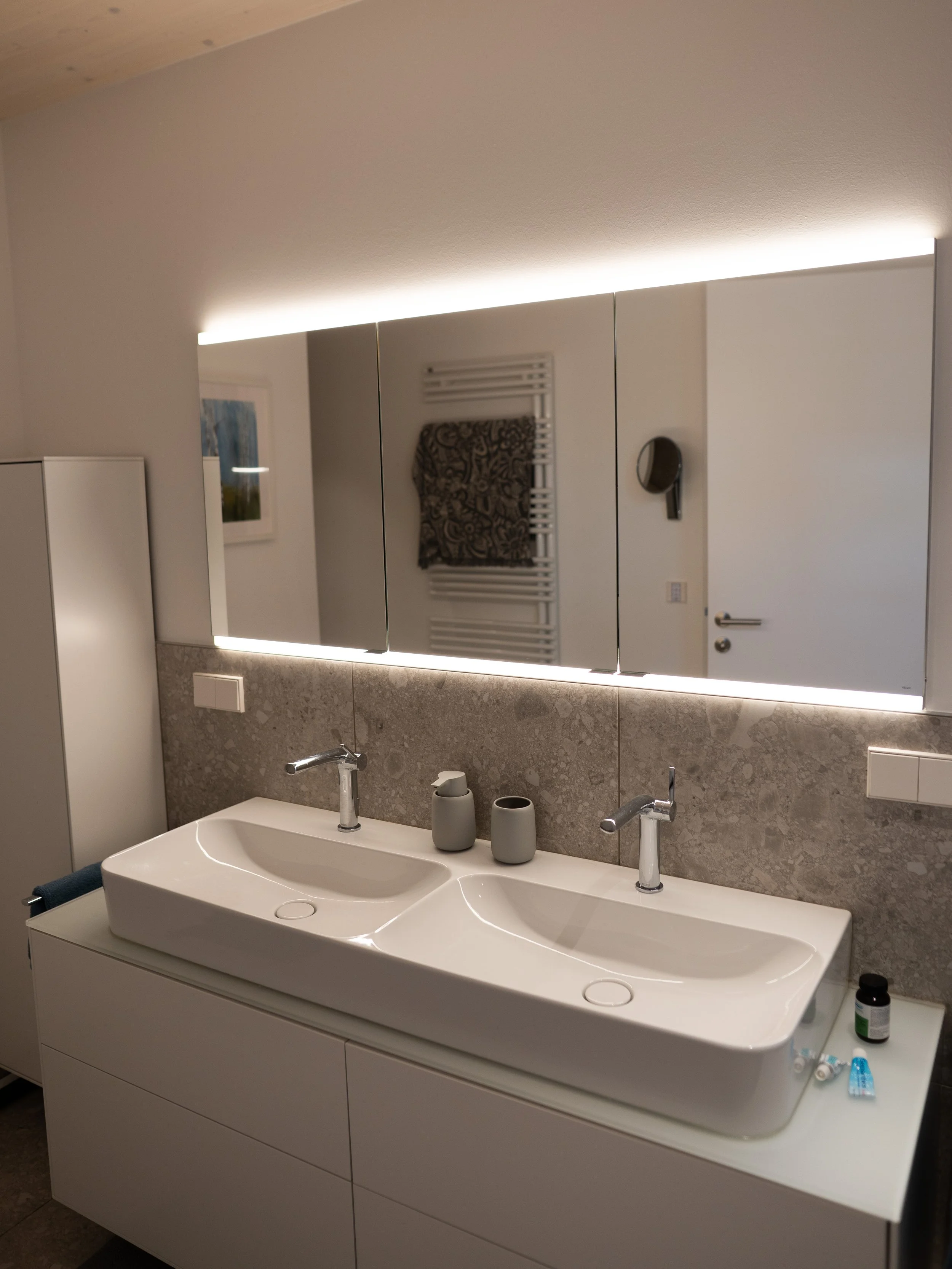 Ein Badezimmer mit Doppelwaschbecken, Spiegel mit LED-Beleuchtung, Handtuchhalter mit Handtuch, Wasserhähnen, zwei grauen Behältern, einer schwarzen Tablette, einer kleinen Tube, einem schwarzen Fläschchen und einer weißen Wand mit Heizkörper.