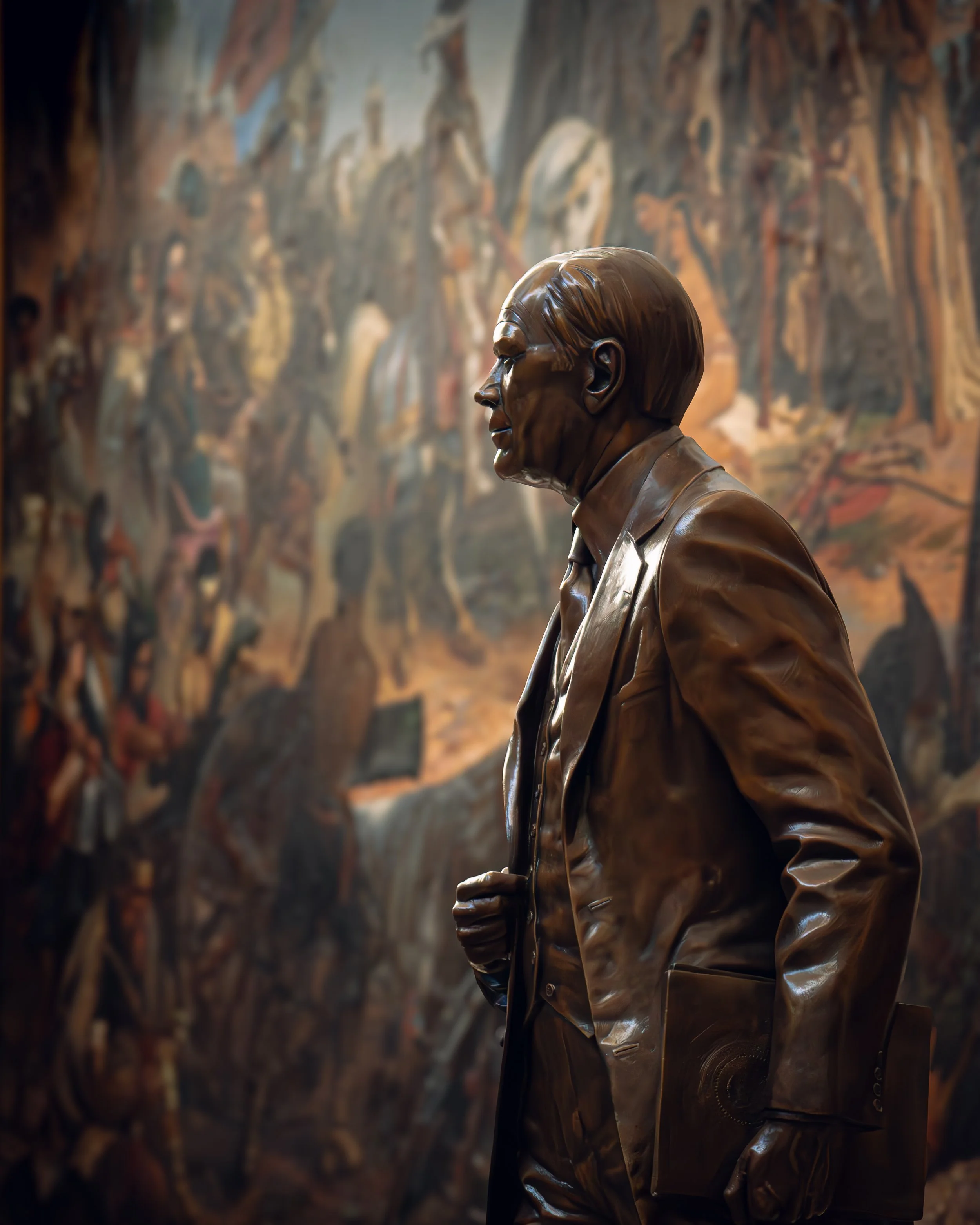 Bronzestatue eines Mannes in einem Anzug, Blick nach links, im Hintergrund Wandgemälde mit zahlreichen Figuren.