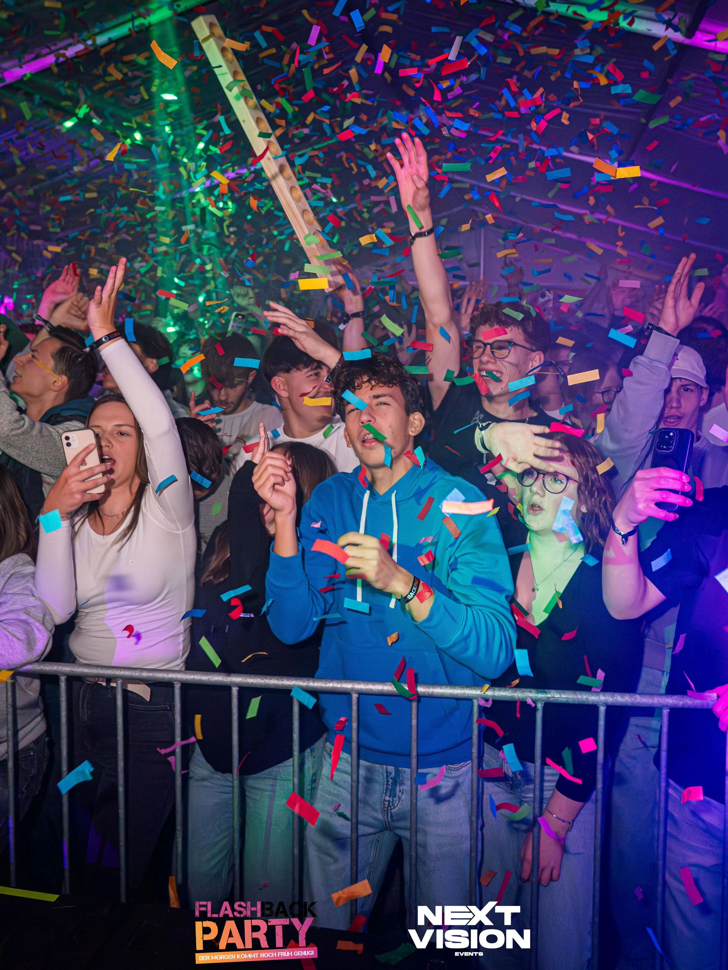 Menschen feiern bei einer Party mit Konfetti, bunte Lichter, einige nehmen Selfies, alle scheinen Freude zu haben.