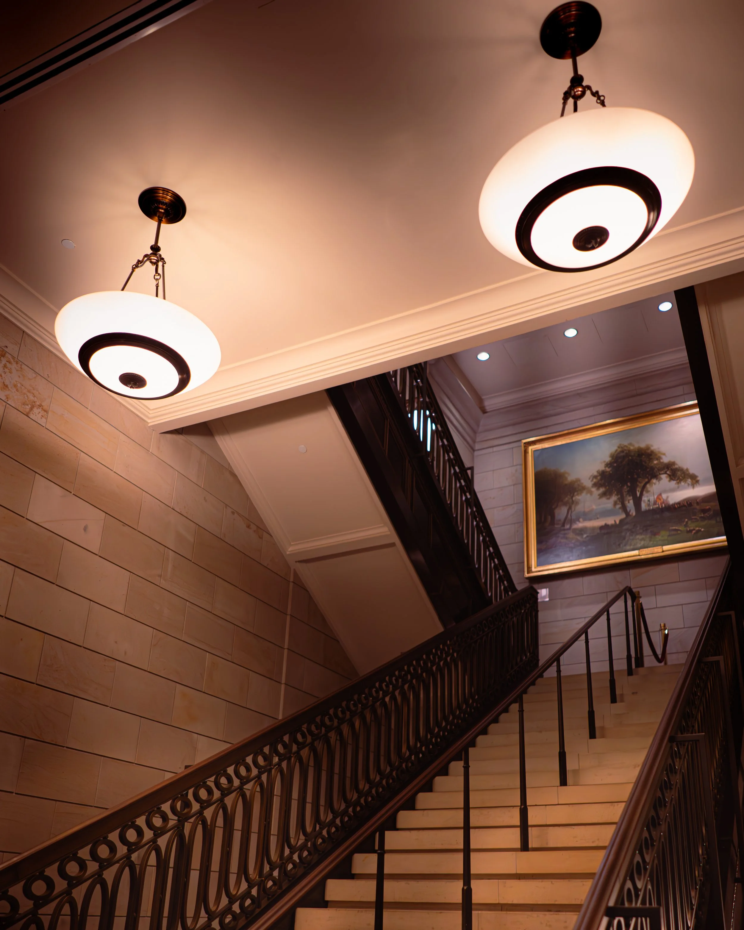 Treppenhaus mit two Deckenleuchten, Wandbild mit Landschaftsmotiv, breite Treppe mit verzierten Geländern, Innenraum mit eleganter Dekoration.