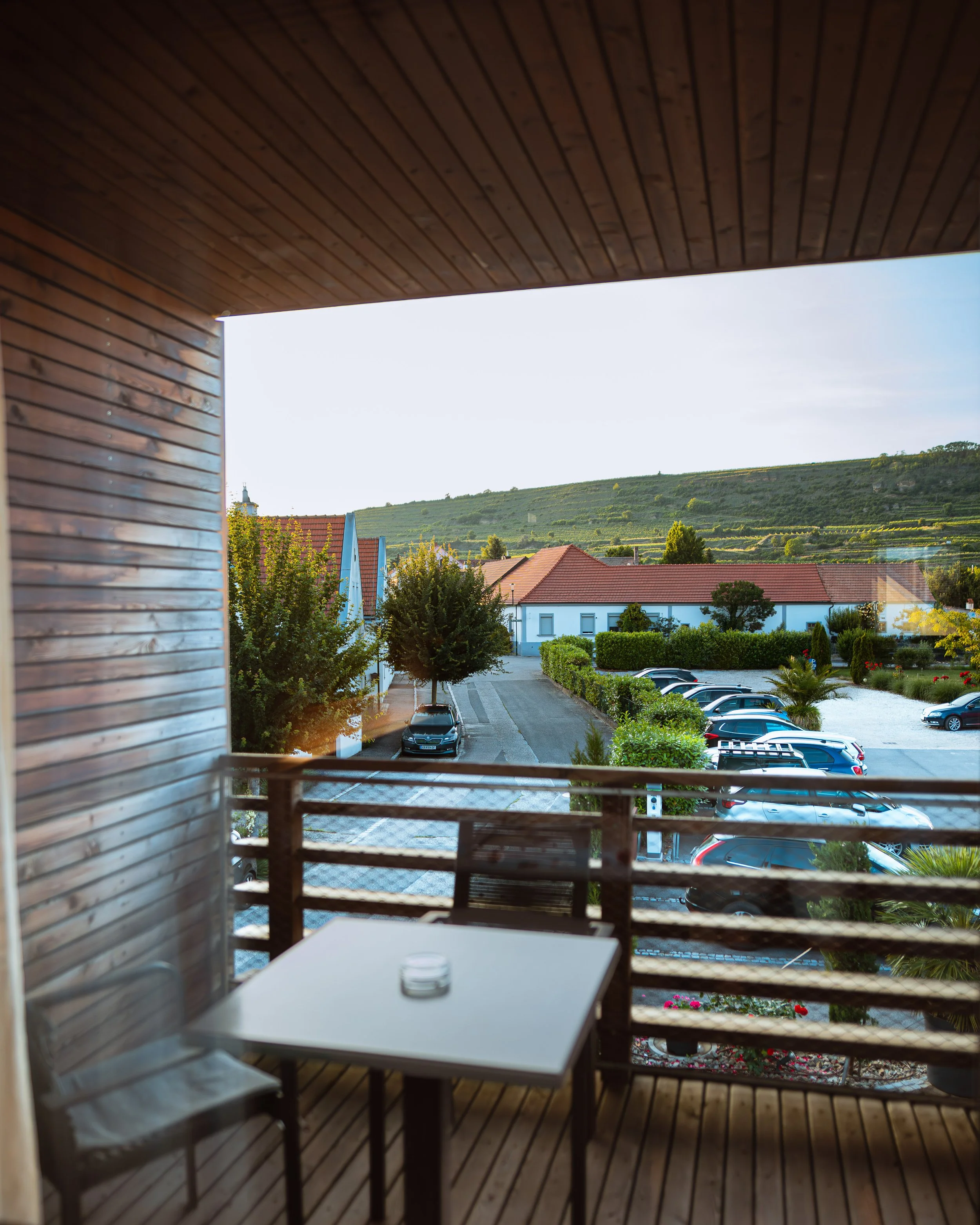 Blick von einem Balkon mit Holzverkleidung auf einen Parkplatz, Bäume und Häuser mit roten Dächern vor einem Hügel im Sonnenlicht.