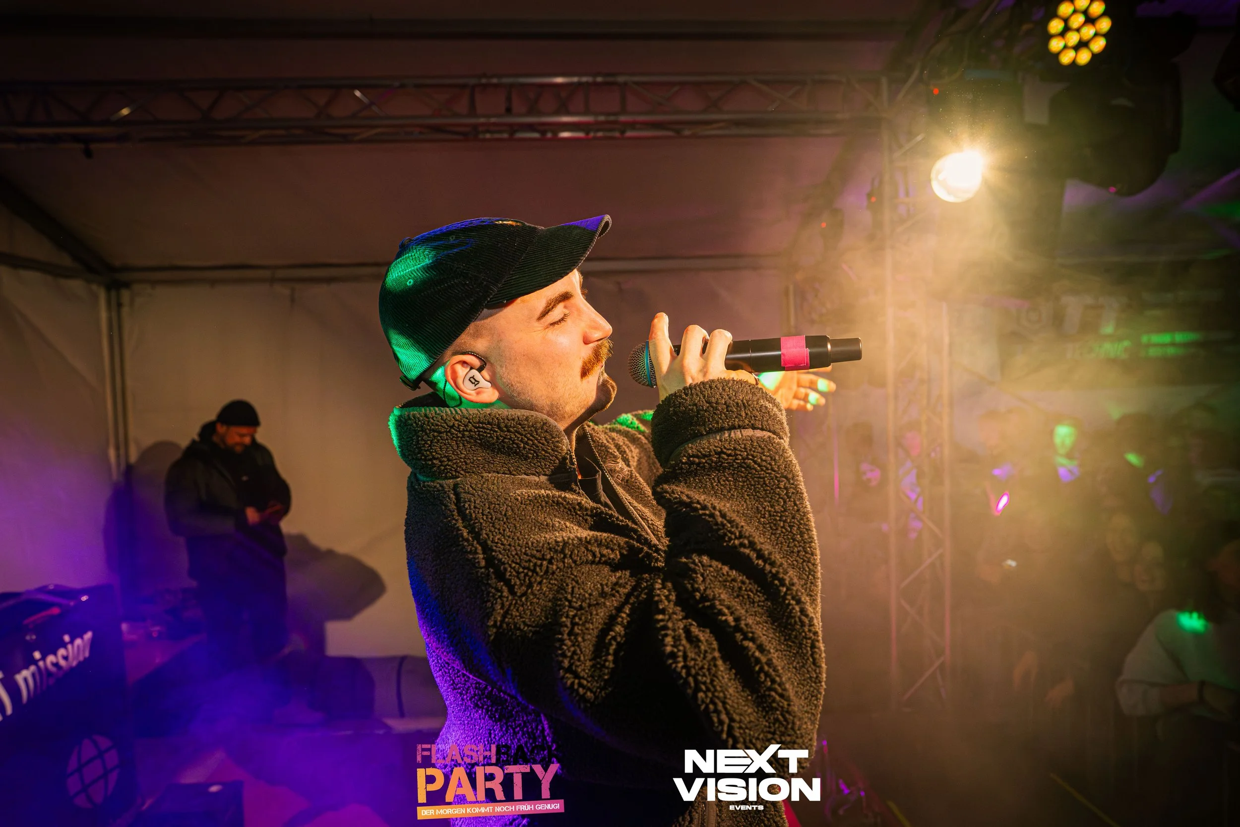 Ein Mann singt mit einem Mikrofon bei einer Party, umgeben von buntem Licht und Rauch, im Hintergrund sind weitere Personen.