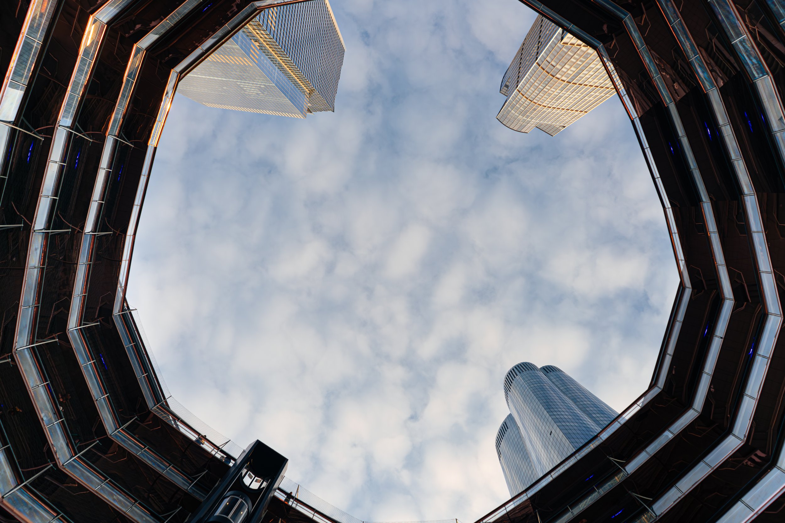 Blick nach oben in einen Innenhof umgeben von modernen Hochhäusern mit Glasfassaden, die den Himmel spiegeln.