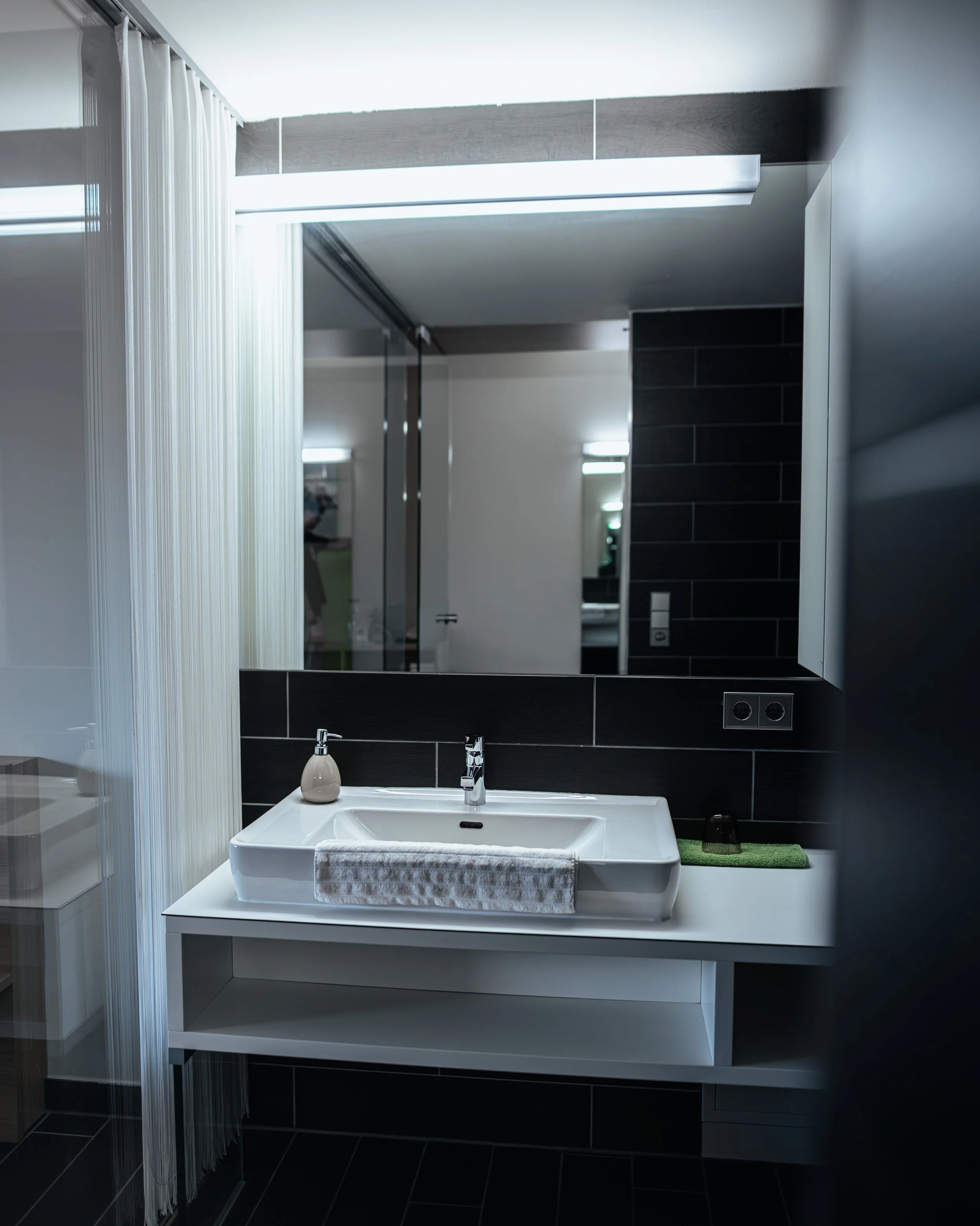 Moderner Waschtisch mit Spiegel, dunkler Fliesenwand und schwarzen Steckdosen in einem Badezimmer.