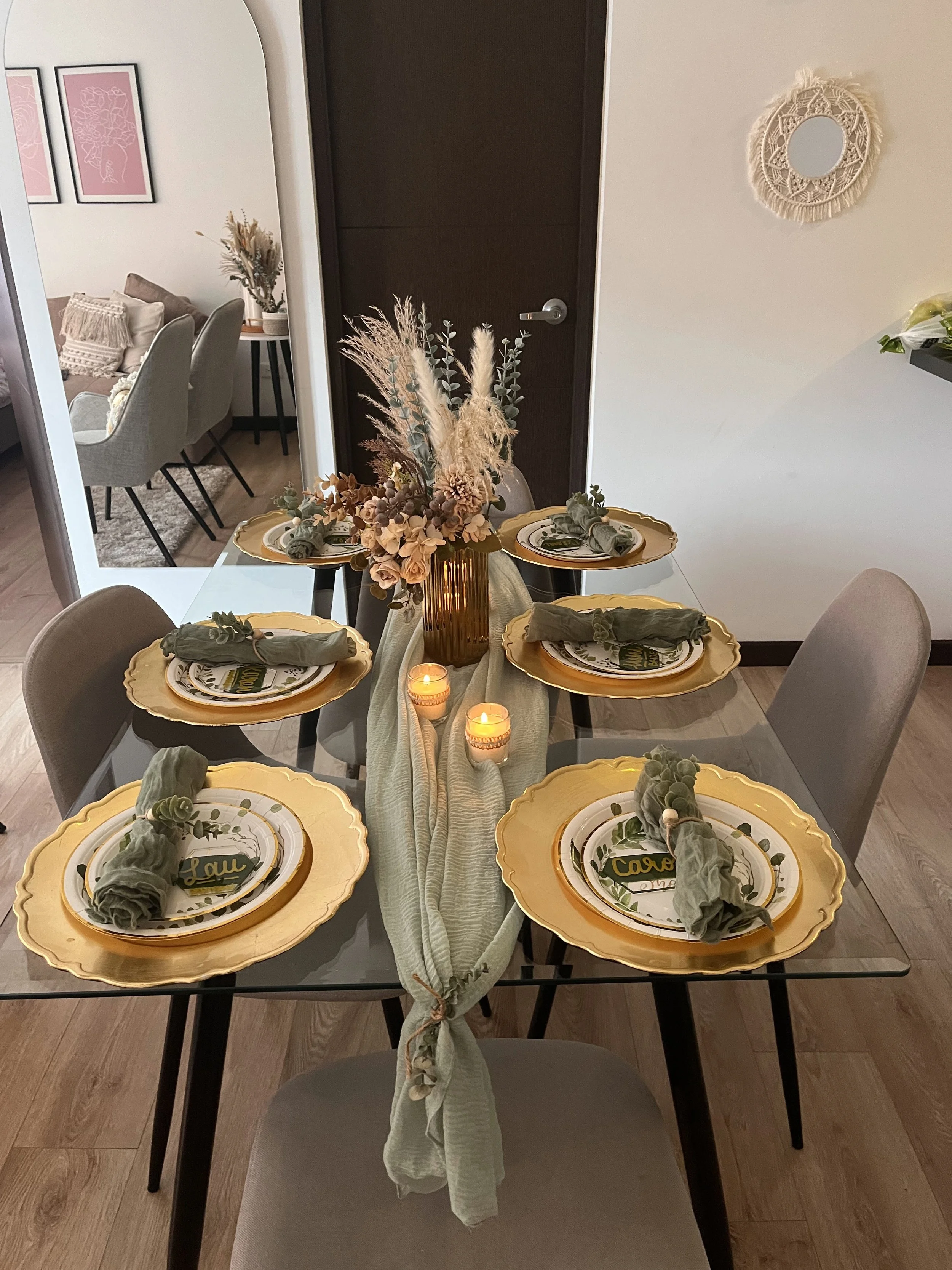 A dining table set for six with yellow scalloped plates, green napkins, and decorative place cards, with a centerpiece of dried flowers in a brown vase and lit candles, in a modern dining room.