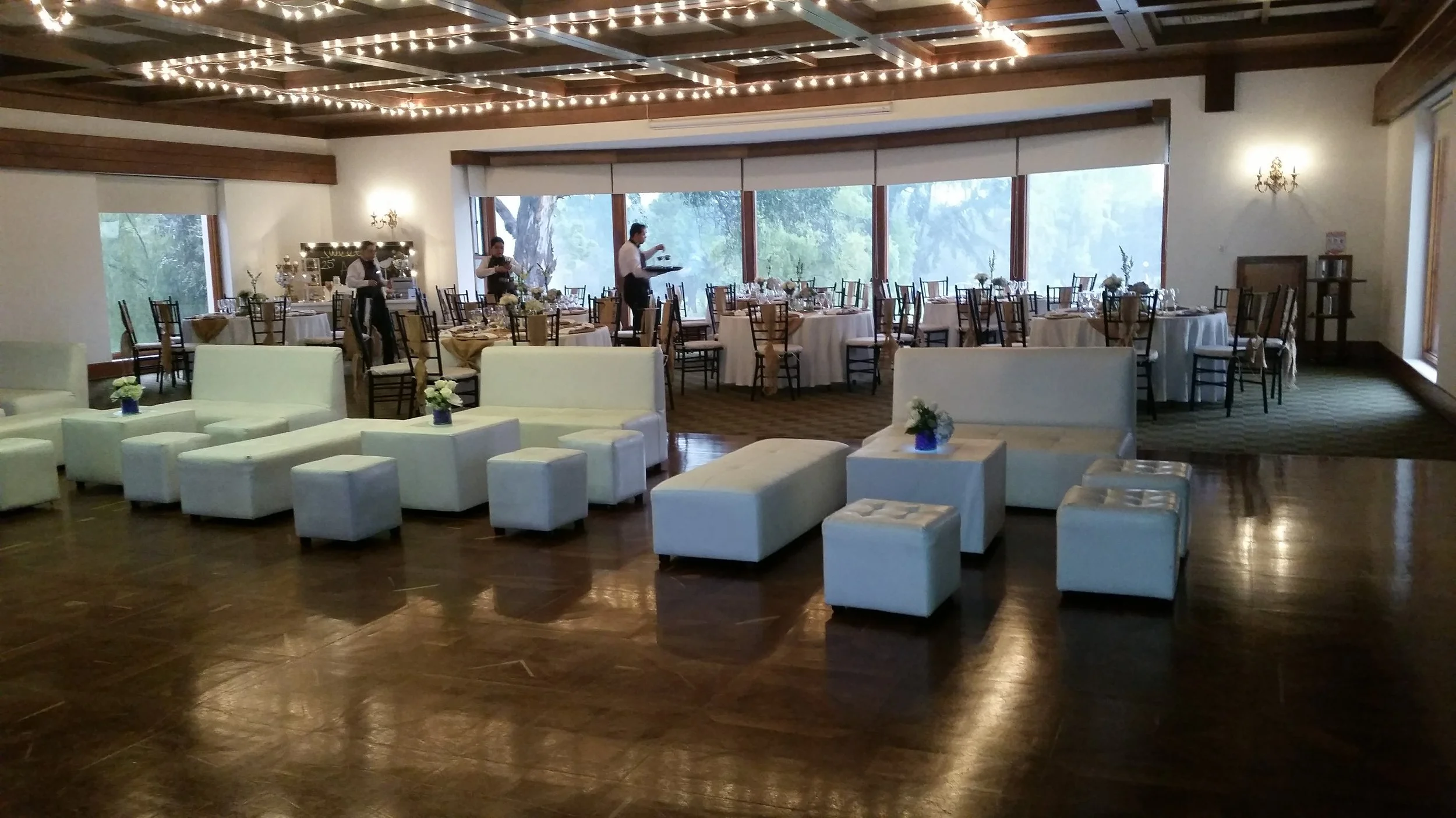 Elegant banquet hall with round tables set for a formal event, decorated with white flowers, and a seating area with white sofas and ottomans in the foreground. Waitstaff are preparing the tables near large windows with greenery outside, and string lights are hanging from the wooden ceiling.