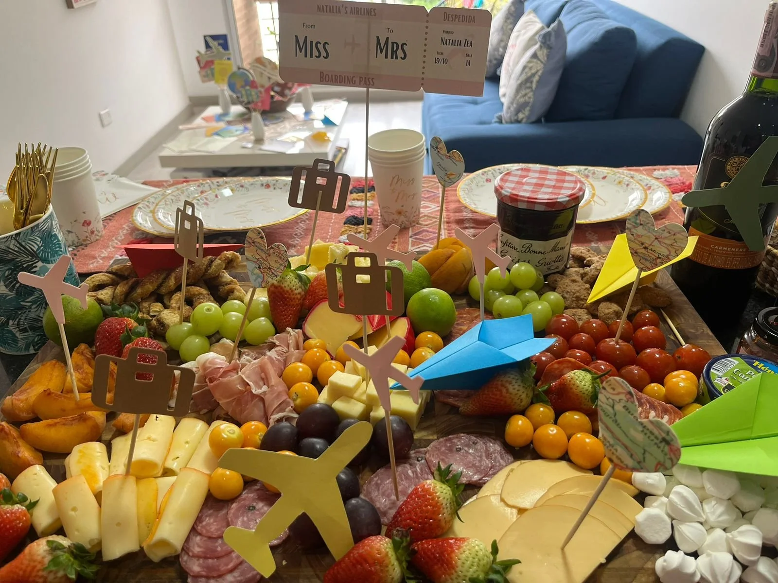 A festive spread of fruit, cheese, and charcuterie on a table, decorated with colorful paper airplanes and travel-themed paper flags, set for a celebration or gathering.