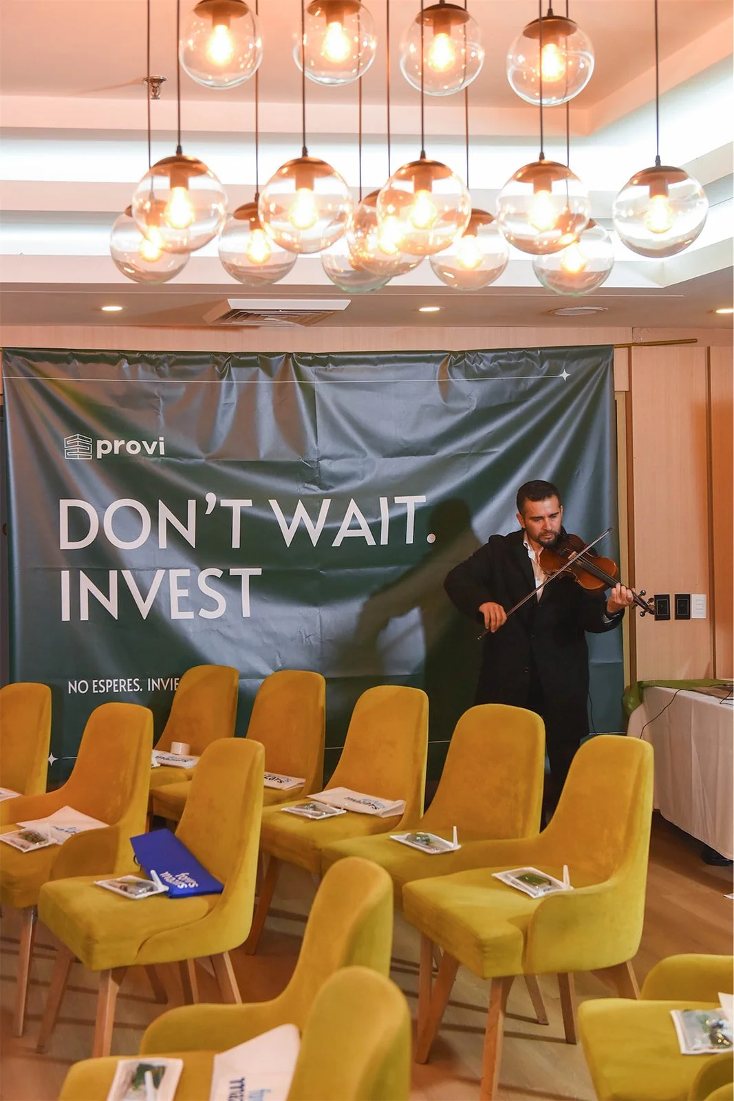 Hombre tocando violín en una sala con sillas amarillas, cartel en el fondo que dice 'Don't Wait. Invest' y decoración moderna con lámparas colgantes.