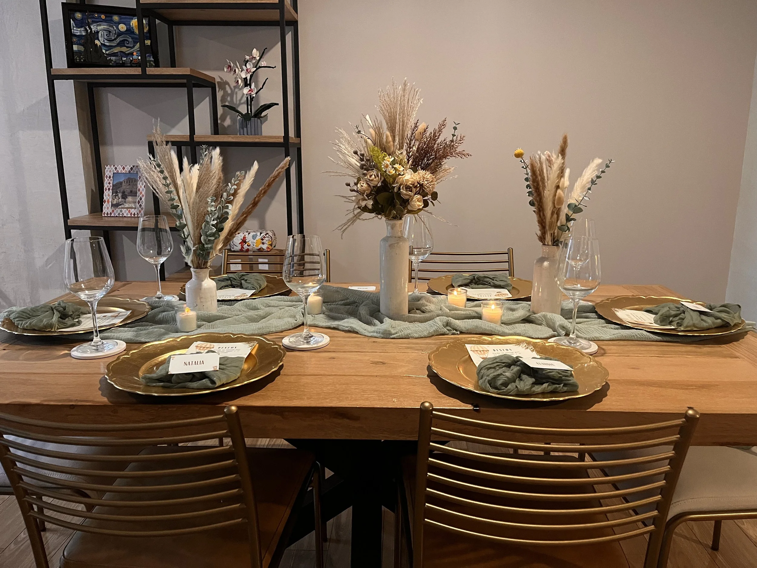 A decorated dining table set for a meal with six place settings. The table features gold charger plates, green napkins, and small card place cards. A green table runner, candles, and floral centerpieces with dried grasses and flowers are arranged in vases. In the background, a black metal shelving unit holds decorative items and framed photos.
