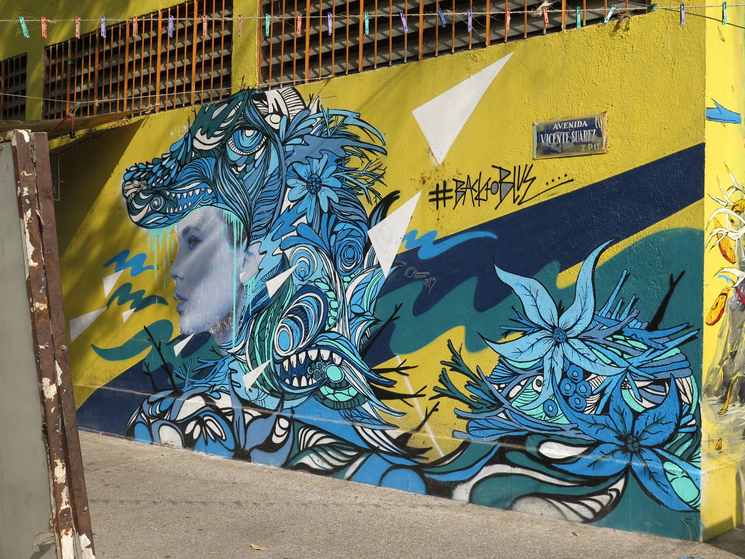 Colorful street art mural on a yellow wall features a woman's profile with flowing, intricate blue and black decorative patterns, flowers, and a large bird in the background.
