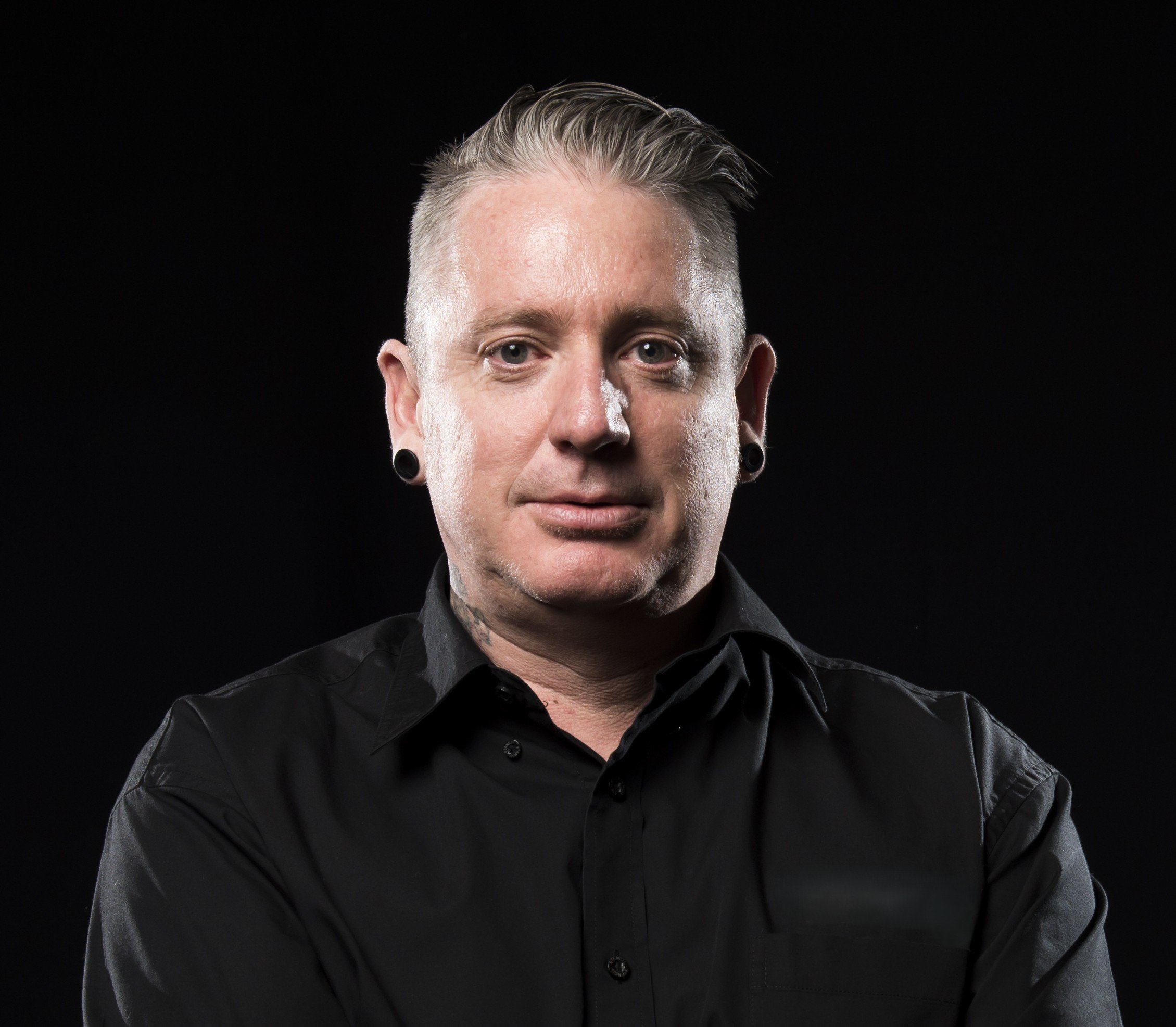 Portrait of a man with platinum blonde hair, wearing a black shirt with rolled-up sleeves, earrings, and a tattoo on his neck, against a black background.