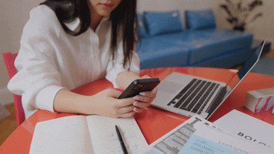 Person in white shirt using smartphone at a desk with a laptop, papers, and a notebook.