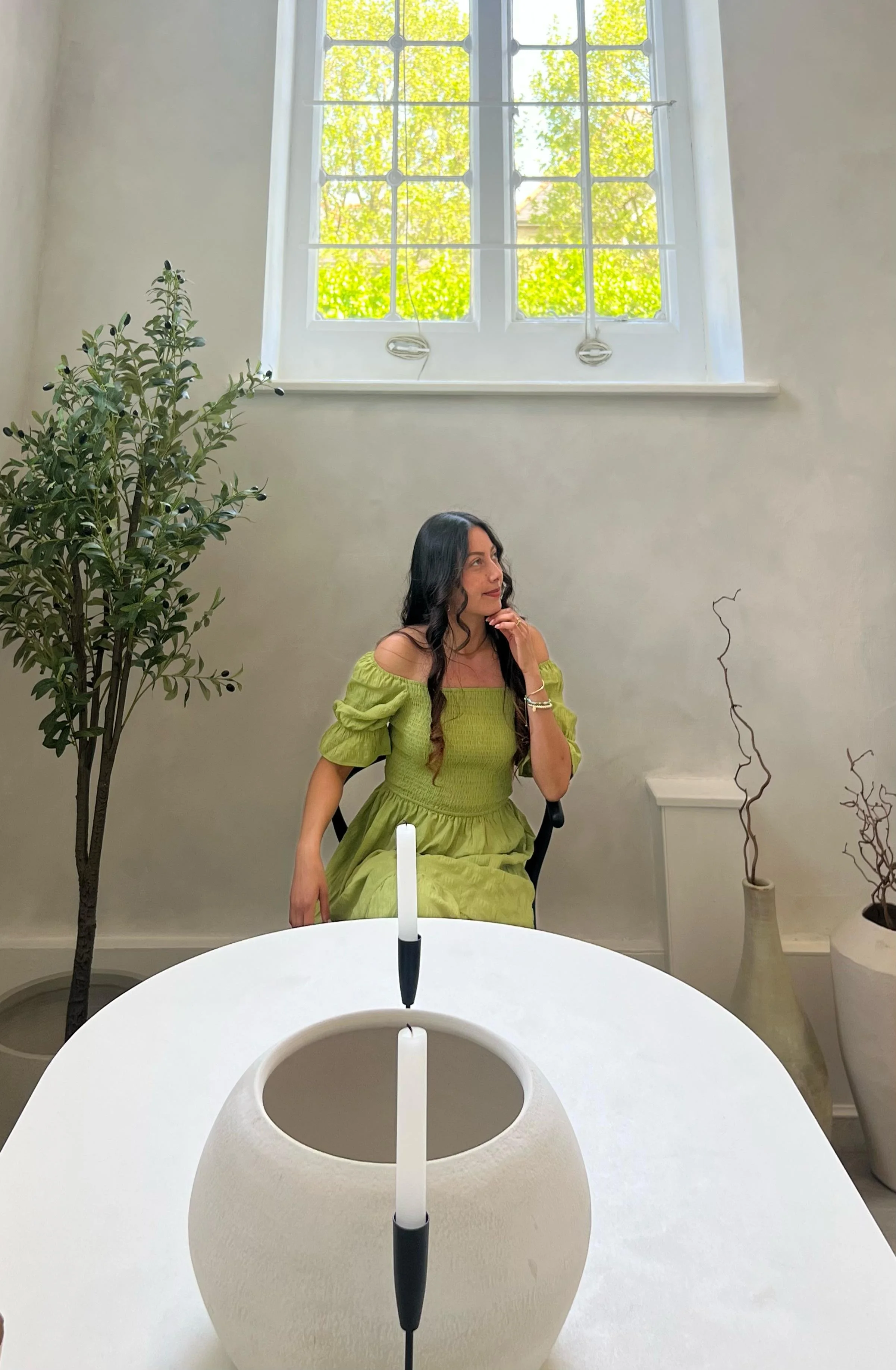 A woman in a light green dress sitting at a white table with two candlesticks, next to a potted plant, in a room with a large window.