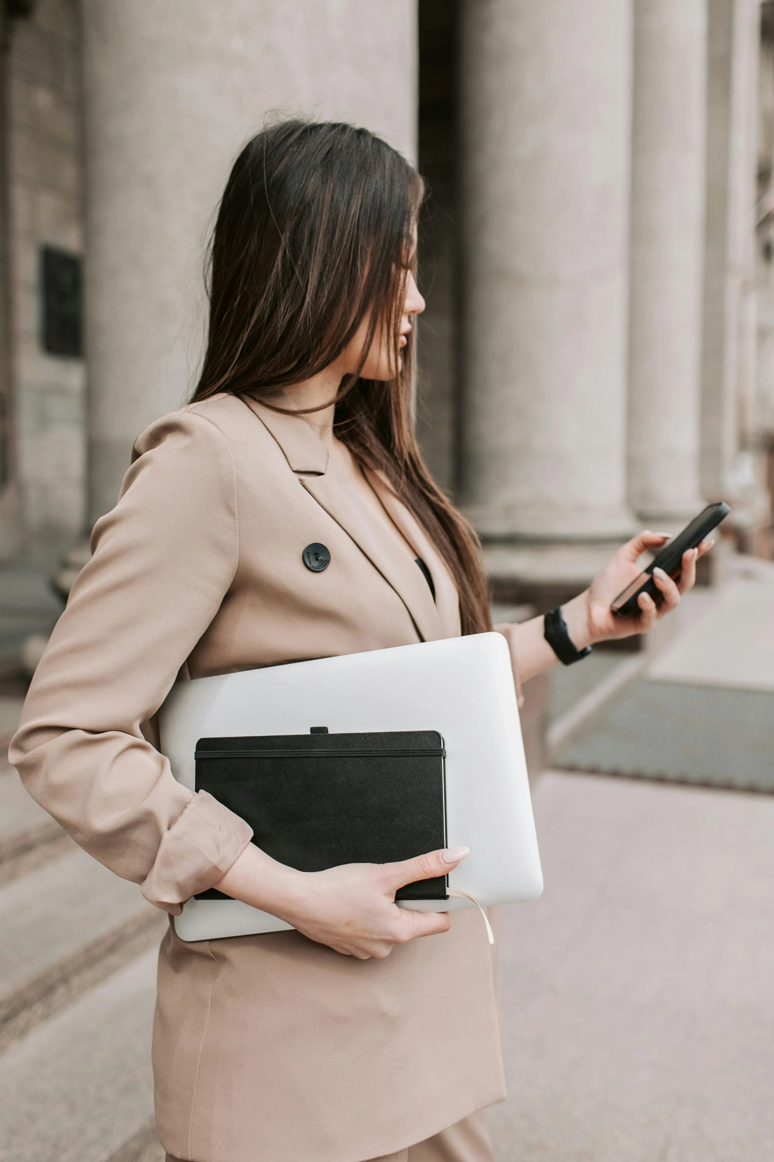 Woman in beige blazer holding a laptop and notebook, using a smartphone, standing in front of building columns.