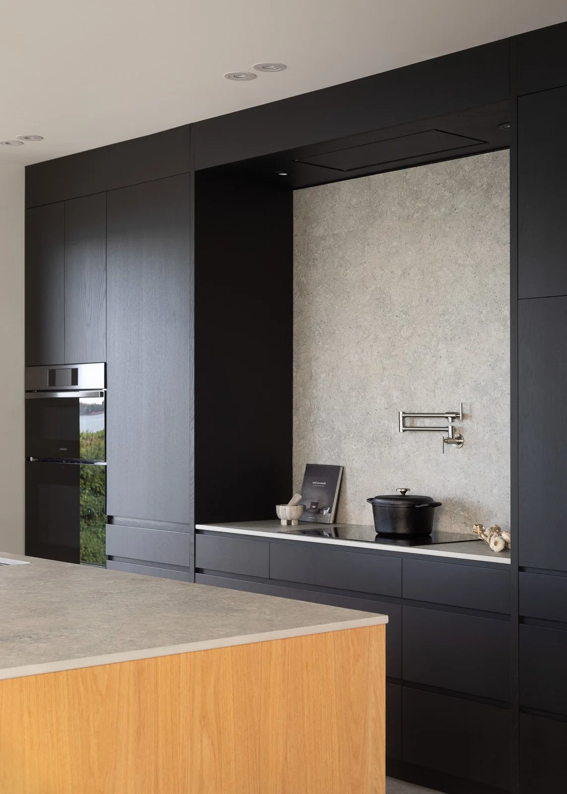 Cuisine moderne noire avec îlot central en bois, plaque de cuisson, hotte intégrée et four encastré.