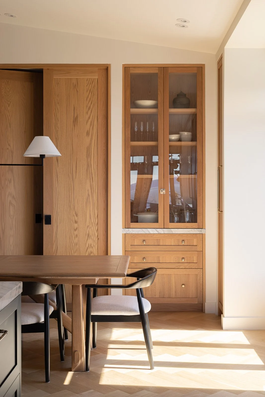 Salle à manger moderne avec table en bois, chaises noires, vitrine intégrée et vaisselier en bois éclairé par la lumière naturelle.