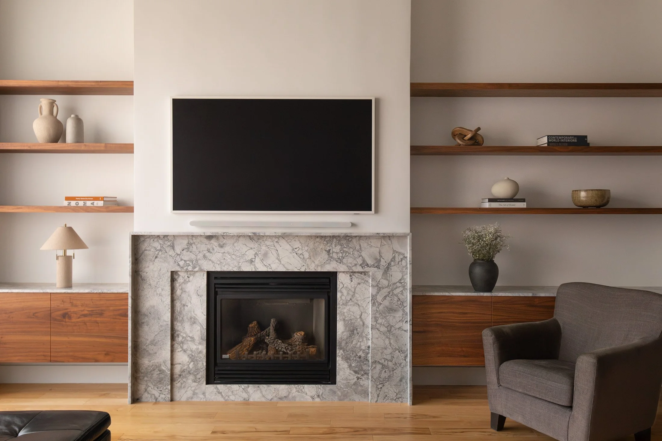 Salon moderne avec cheminée en marbre, téléviseur mural, étagères en bois décorées et fauteuil gris.