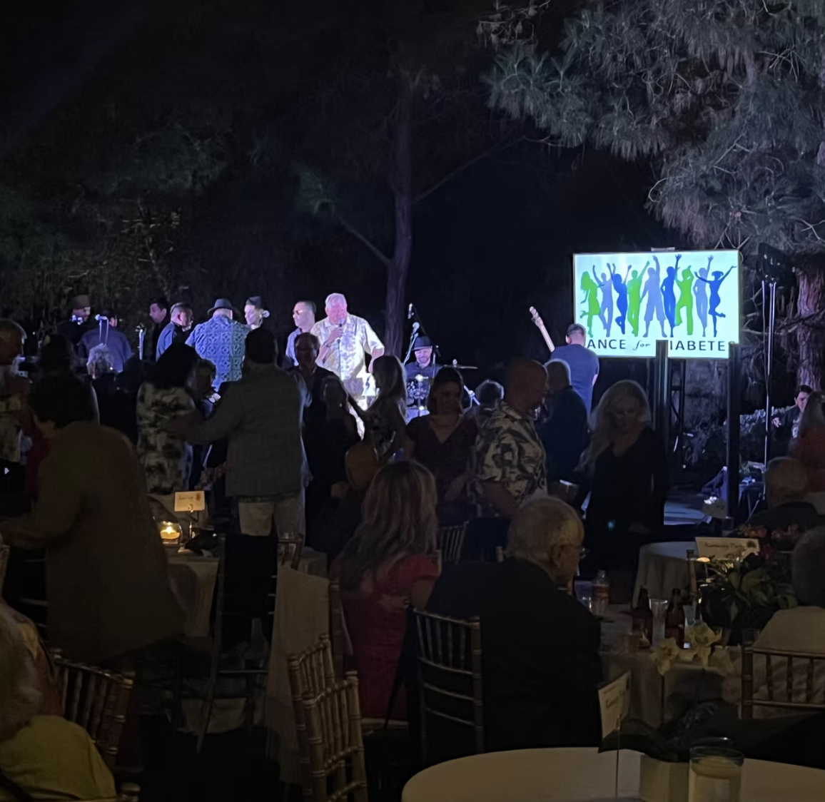 Evening event with people dancing and socializing, live band on stage, illuminated "Dance for Diabetes" sign.