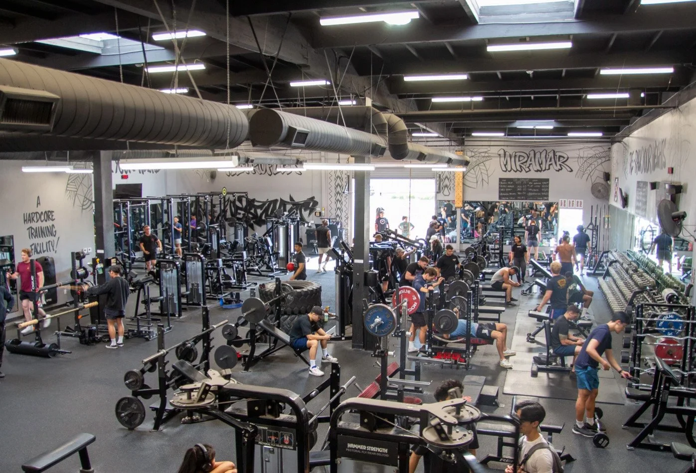 Busy gym with people using various fitness equipment, weightlifting, and exercising in an industrial-style environment.
