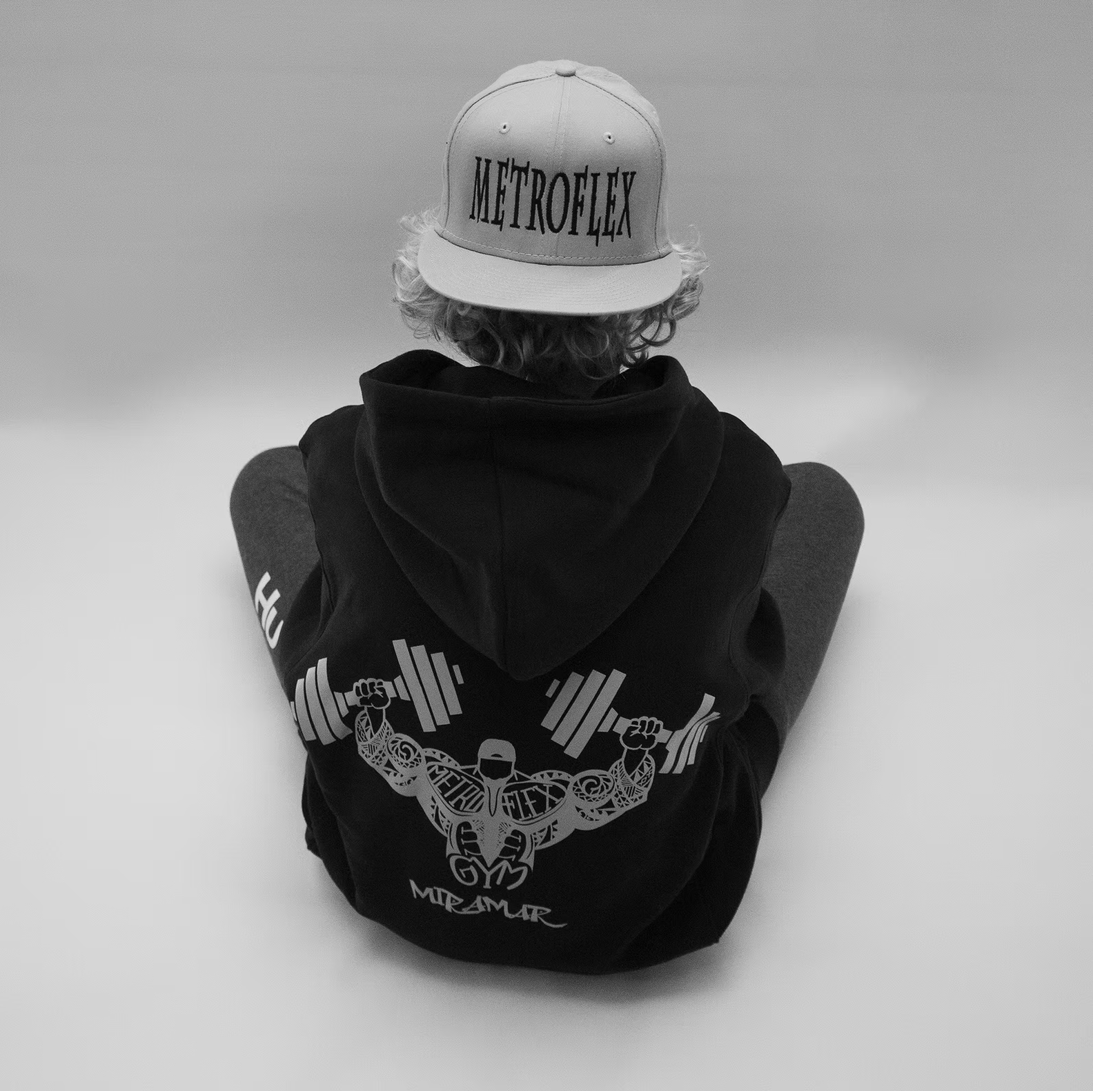 Person wearing a cap with 'Metroflex' and a hoodie featuring a weightlifting graphic, sitting with back to camera.