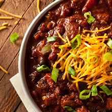 Bowl of chili with grated cheese and green onions topping on a wooden table.