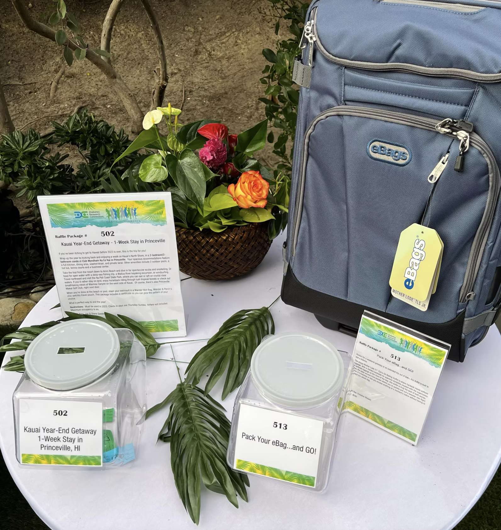 Table setup with a raffle for a Kauai vacation and an eBags luggage package, featuring a blue eBags suitcase, raffle entry boxes, informational signs, tropical decorations, and a floral arrangement.