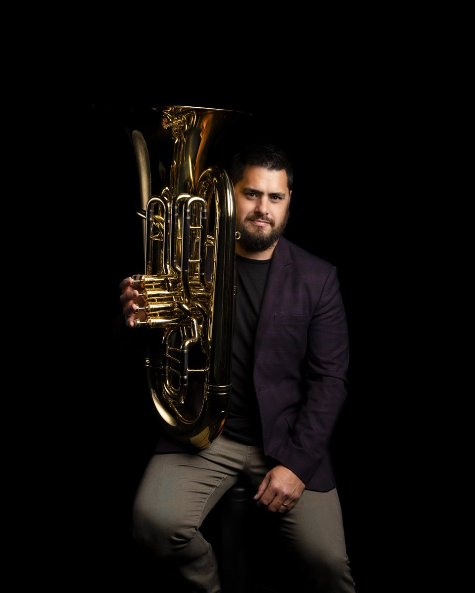 Man with beard and tattoos posing with a brass tuba against a black background.