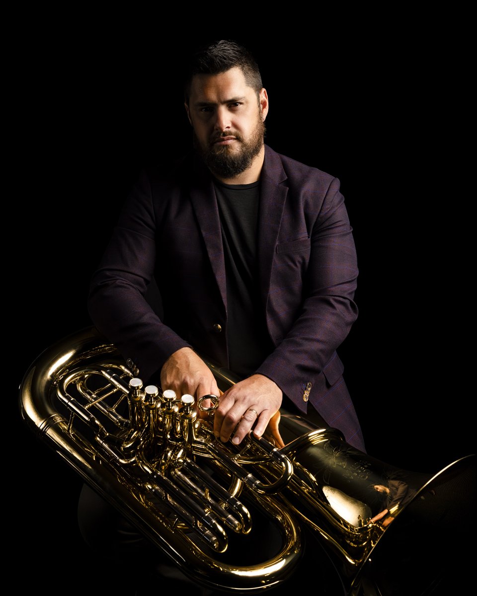 Man with beard and tattoos posing with a brass tuba against a black background.