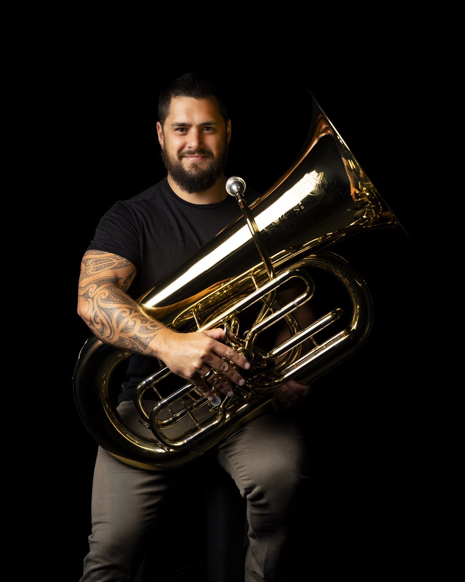Man with beard and tattoos posing with a brass tuba against a black background.