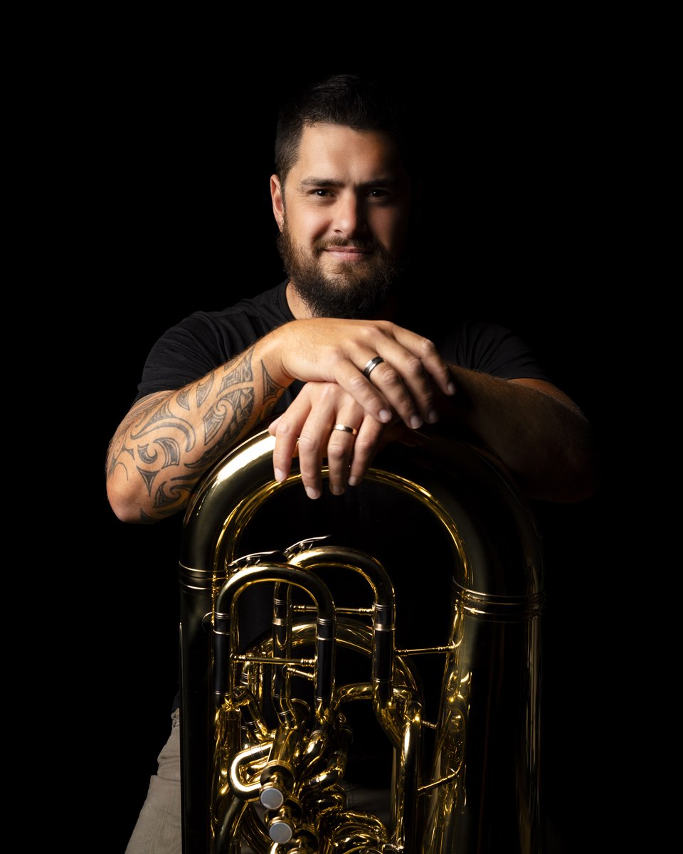 Man with beard and tattoos posing with a brass tuba against a black background.