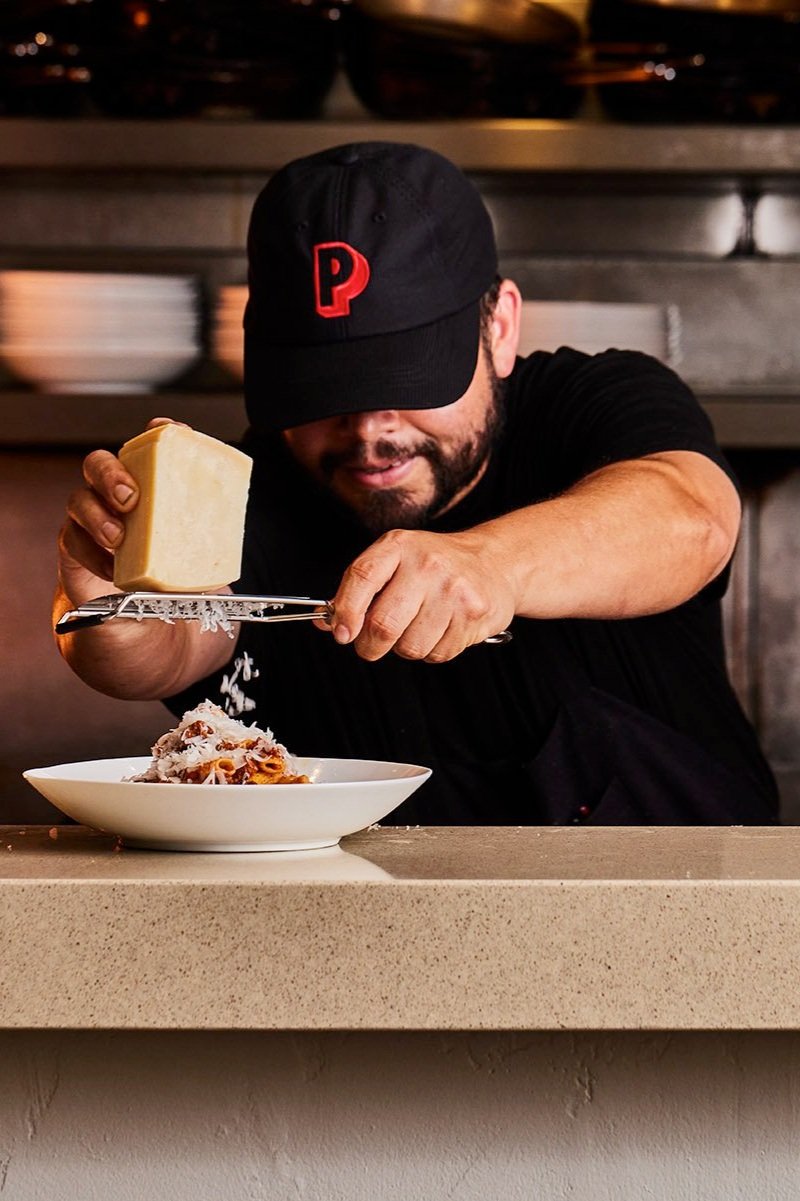 A man in a black cap and shirt sprinkles grated cheese onto a plate of pasta with a cheese grater.