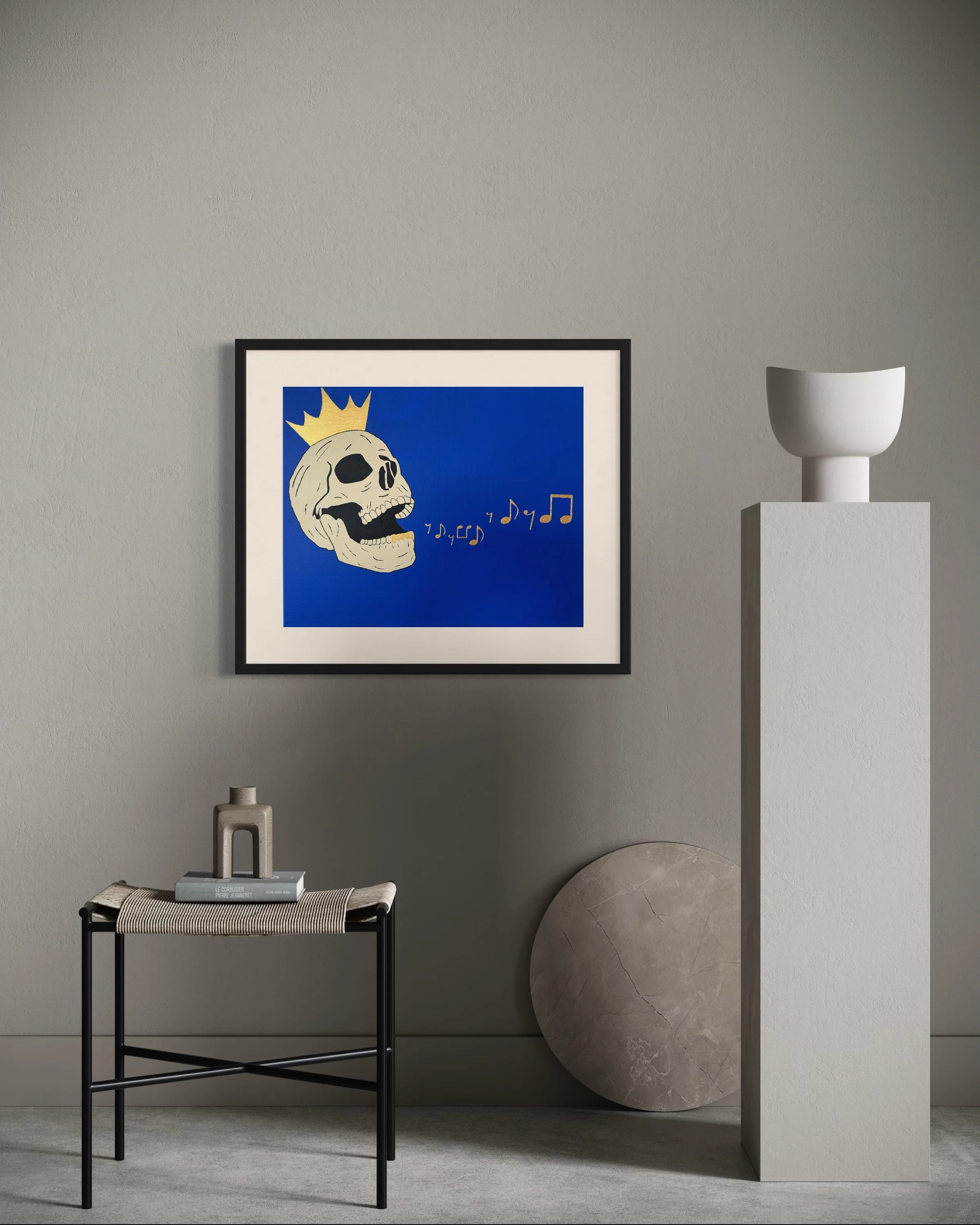 Interior wall with framed artwork of a cartoon skull with a crown, singing musical notes, accompanied by a small stool with decorative objects and a large circular stone object resting against the wall, and a white vase on a pedestal.