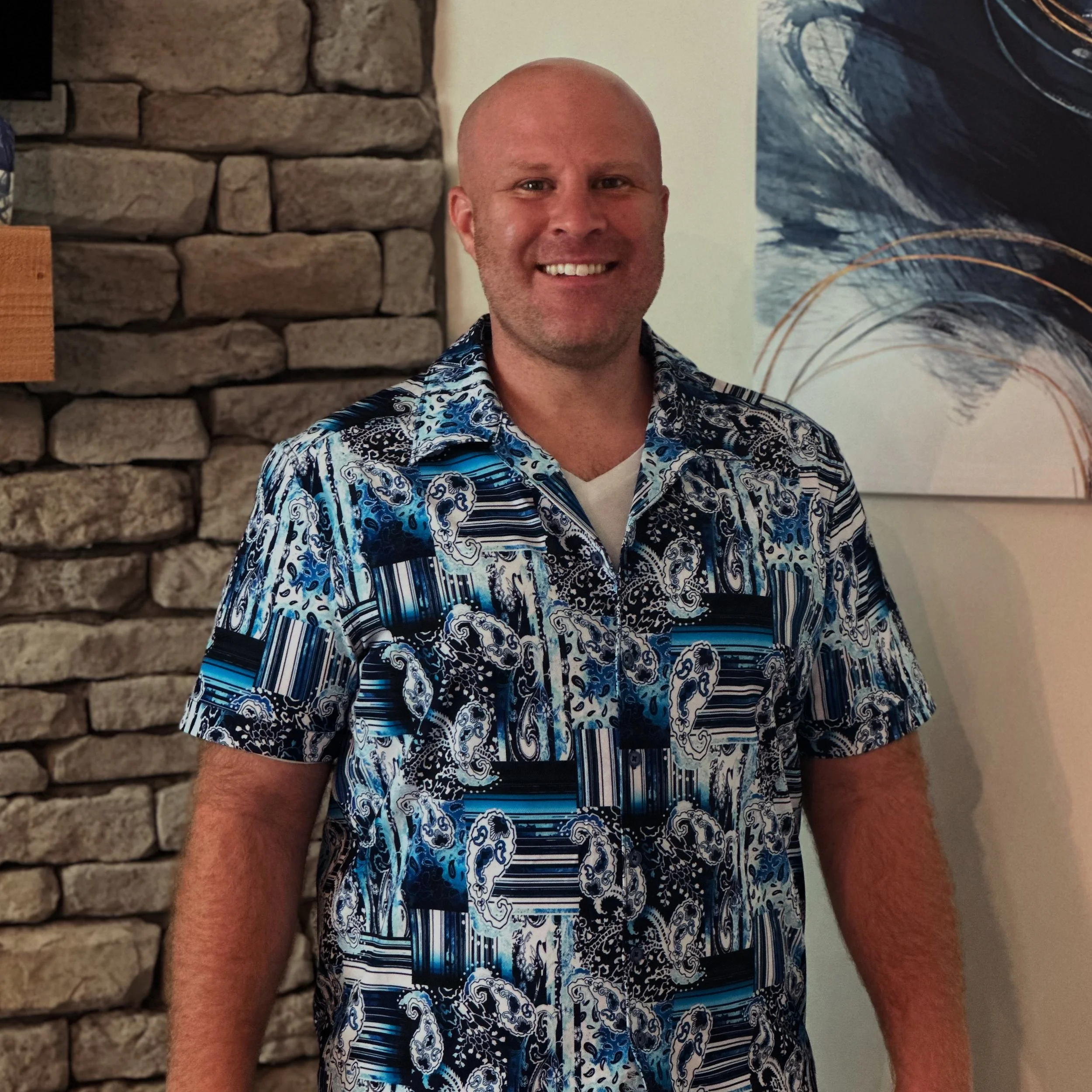 A man with a shaved head smiling, wearing a patterned shirt with blue, black, and white designs, standing indoors with a stone wall and abstract artwork in the background.