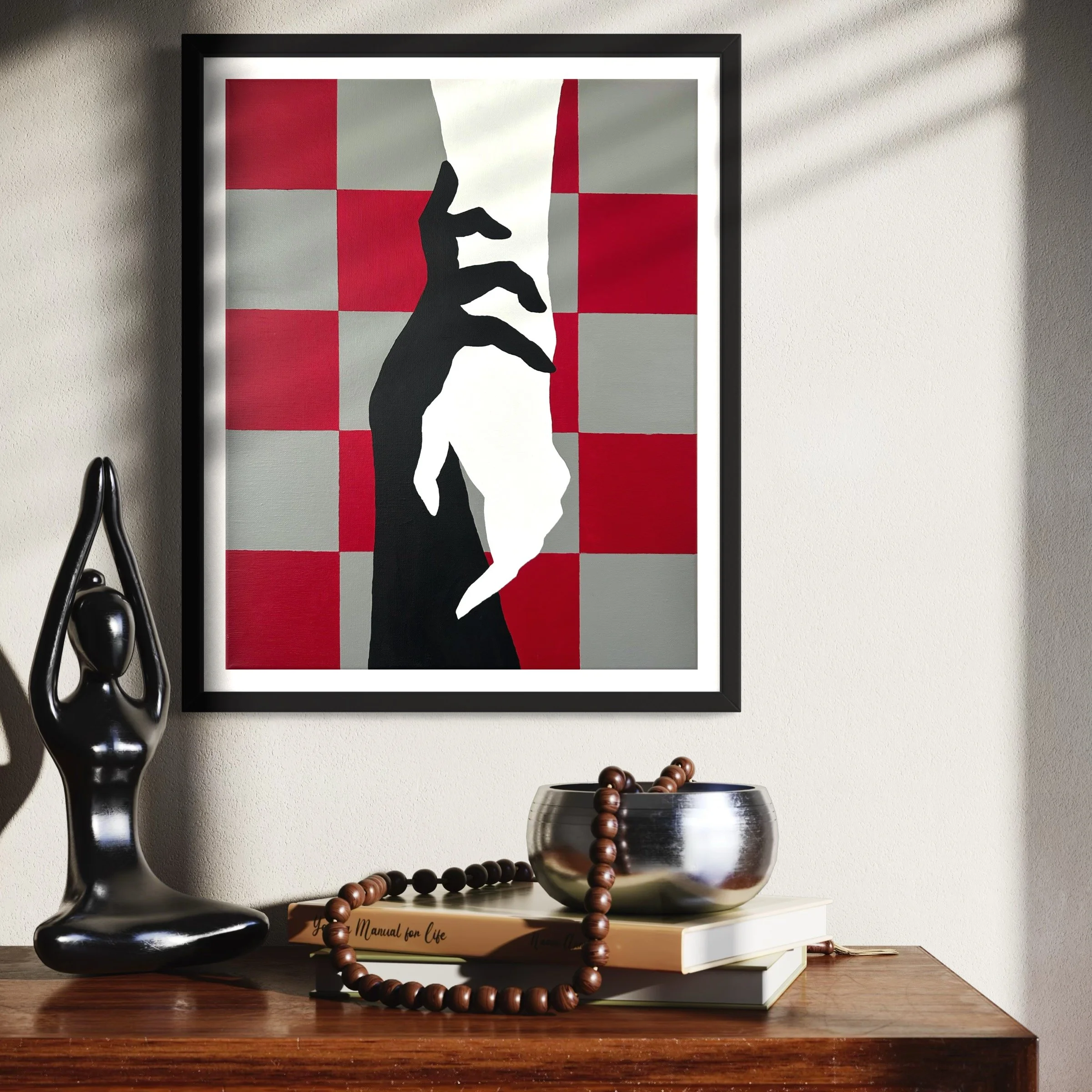 Abstract framed artwork of a black and white hand making a gesture against a red and gray checkerboard background, hanging on a wall above a wooden surface with a black sculpture, a silver bowl with a beaded necklace, and a book.