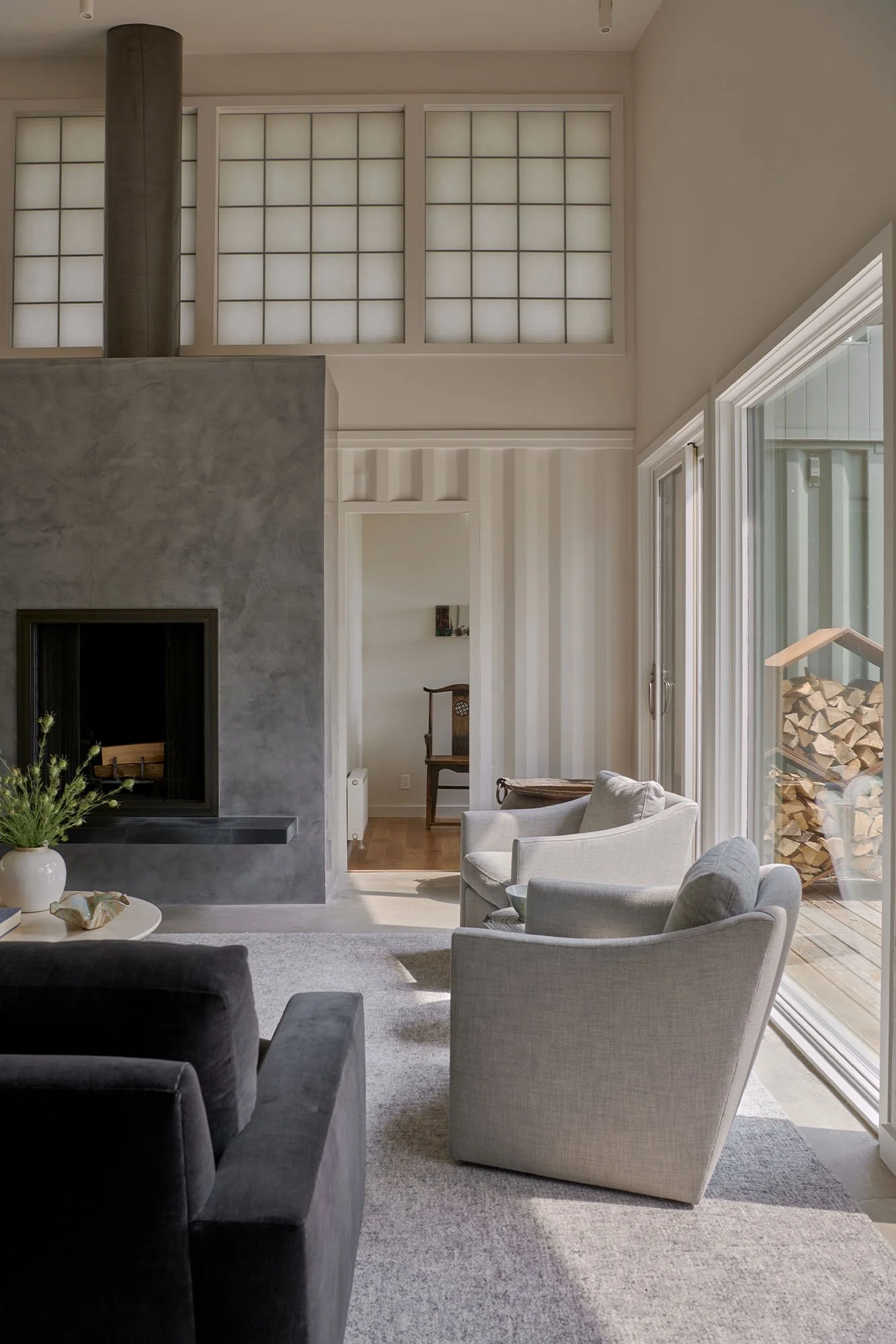 Living room with a gray and black fireplace, white armchairs, sliding glass doors with a woodpile outside, and a doorway leading to another room.