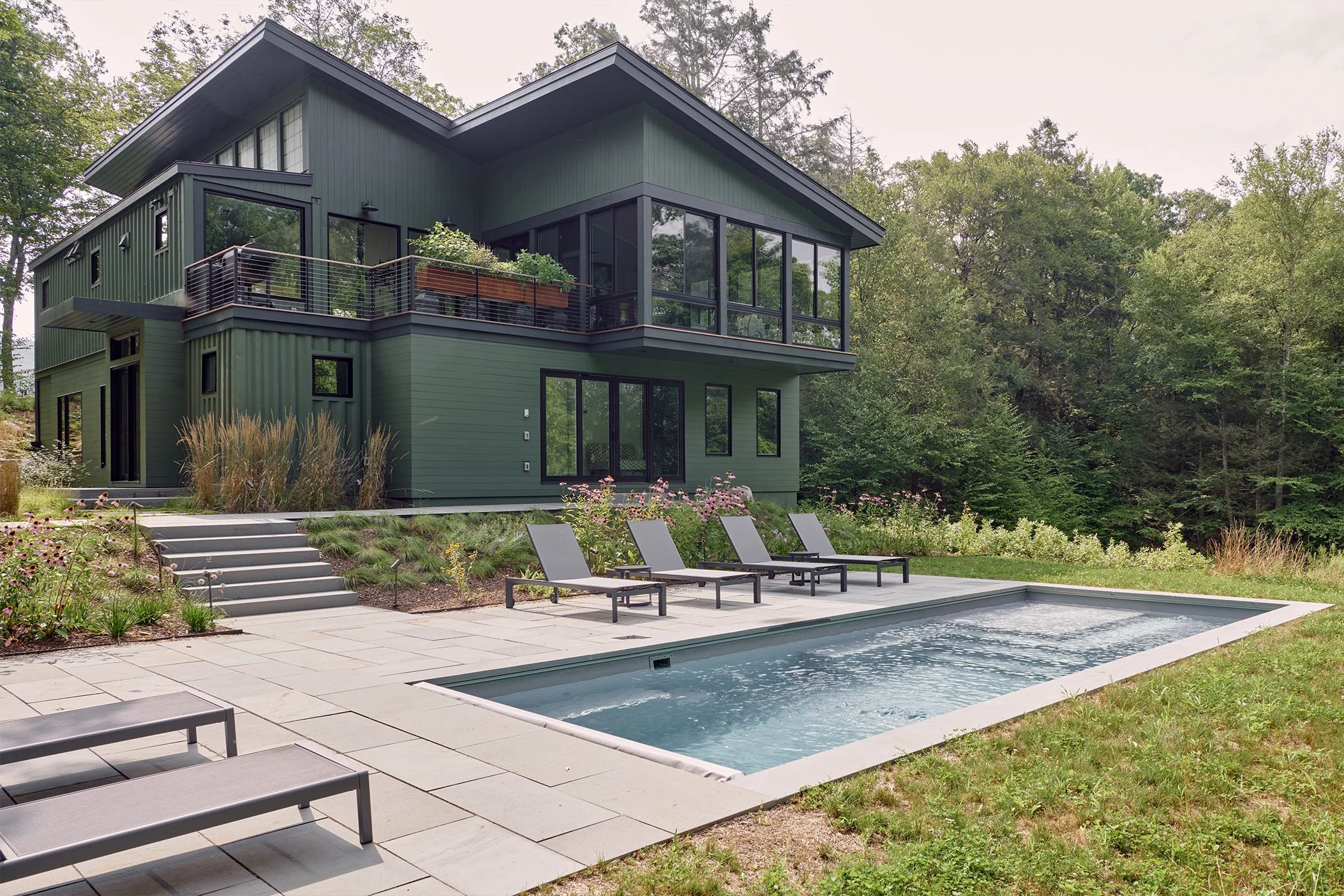 Modern green house with large glass windows, a small pool, and outdoor lounge chairs in a backyard surrounded by trees.