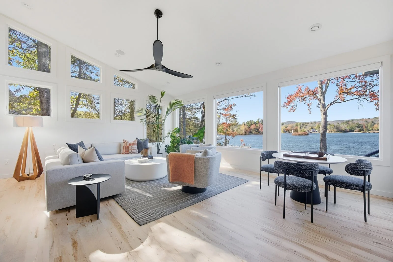 Living room with large windows overlooking a lake, furnished with a white sofa, a round coffee table, a gray armchair with a peach blanket, a black side table, a floor lamp, and dining table with chairs. Contains indoor plants and a ceiling fan.