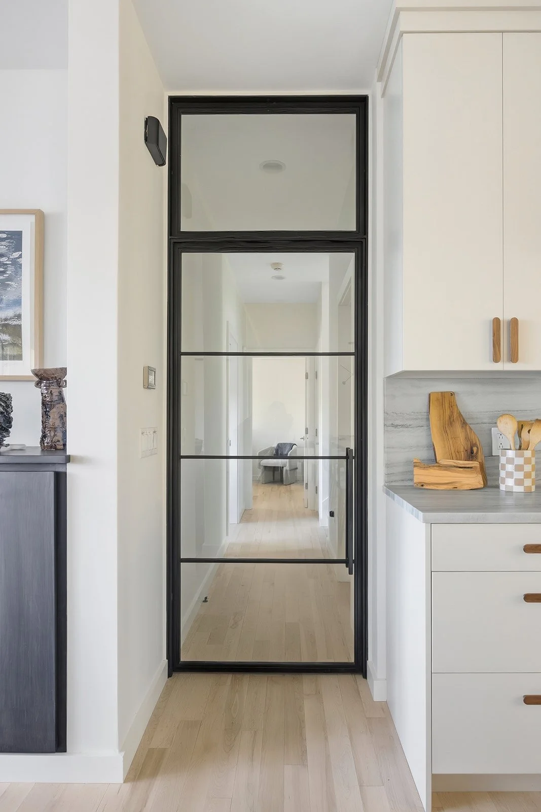 Interior view of a kitchen with a black-framed glass door leading to a hallway with wooden flooring.